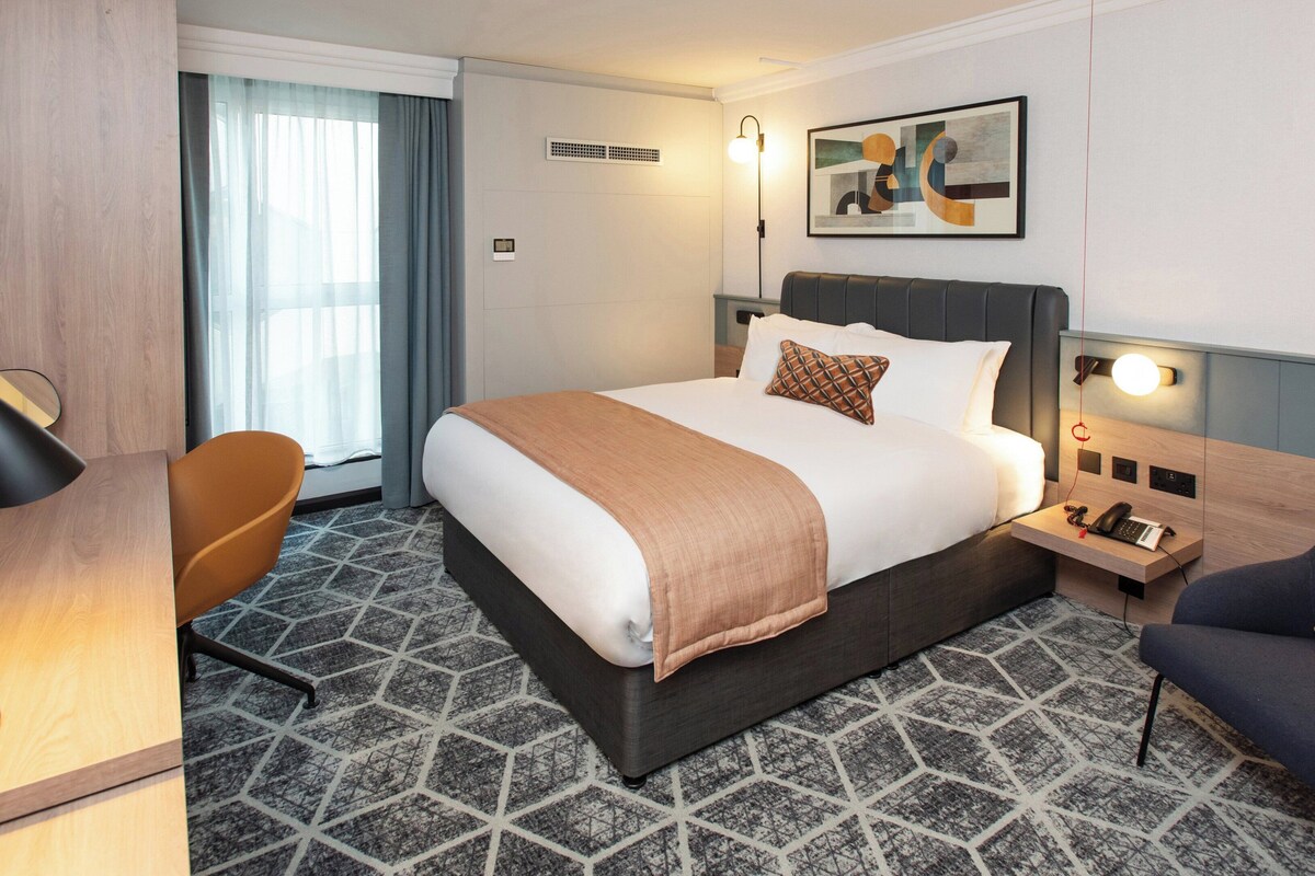 A hotel room with a large bed, a desk with a lamp, a chair, and a bedside table with a phone and a lamp.