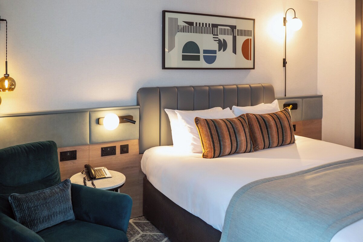 A hotel room with a bed, a chair, a bedside table, and wall-mounted lights.