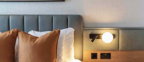 A modern hotel room with a grey upholstered headboard, a round wall-mounted light, and a small round table.