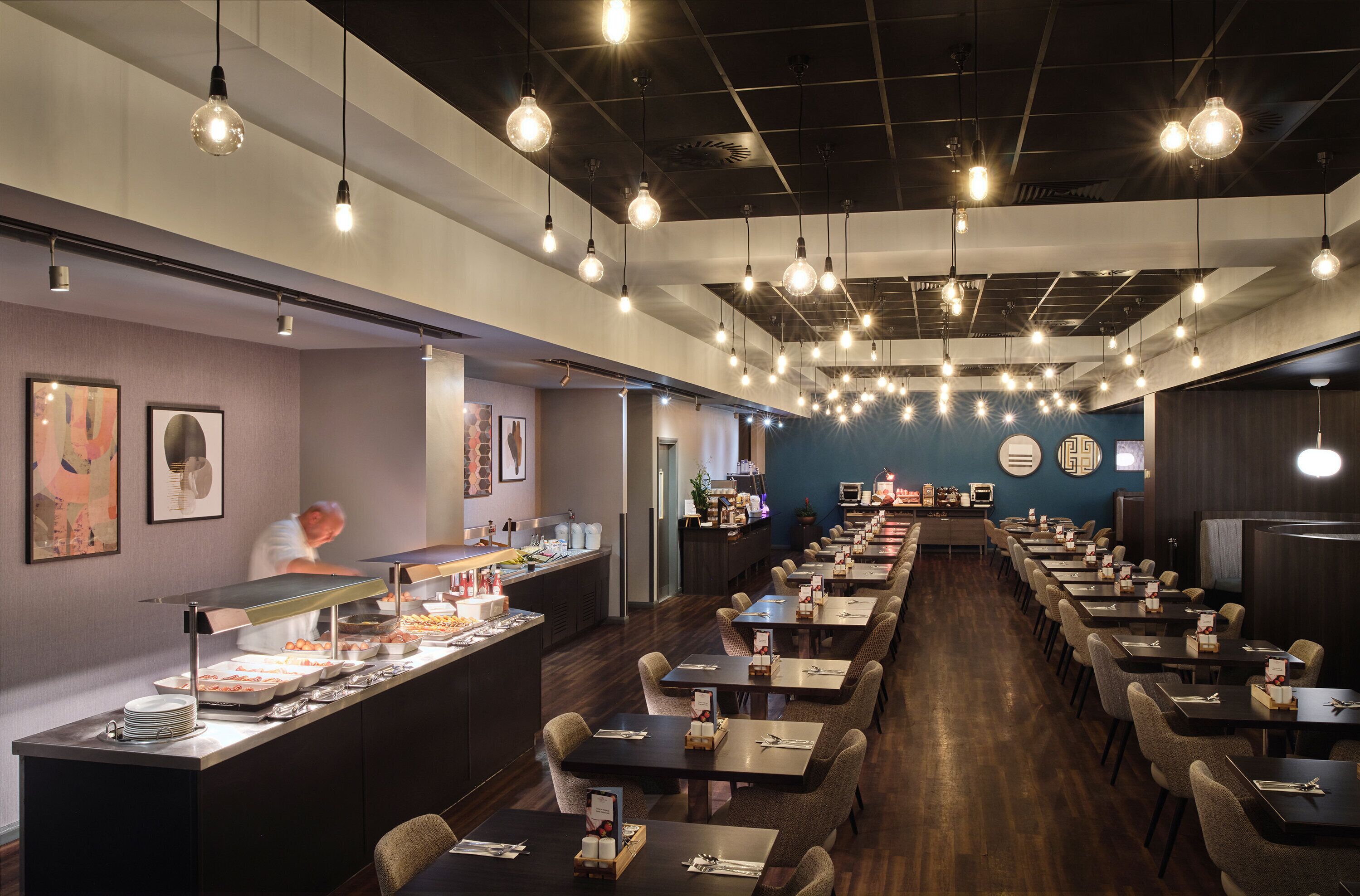A restaurant with a long counter, multiple tables, and hanging light fixtures.