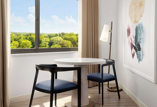 A room with a round table and two chairs, a window with a view of greenery, a lamp, and a piece of abstract art on the wall. - Sonesta White Plains Downtown (White Plains)