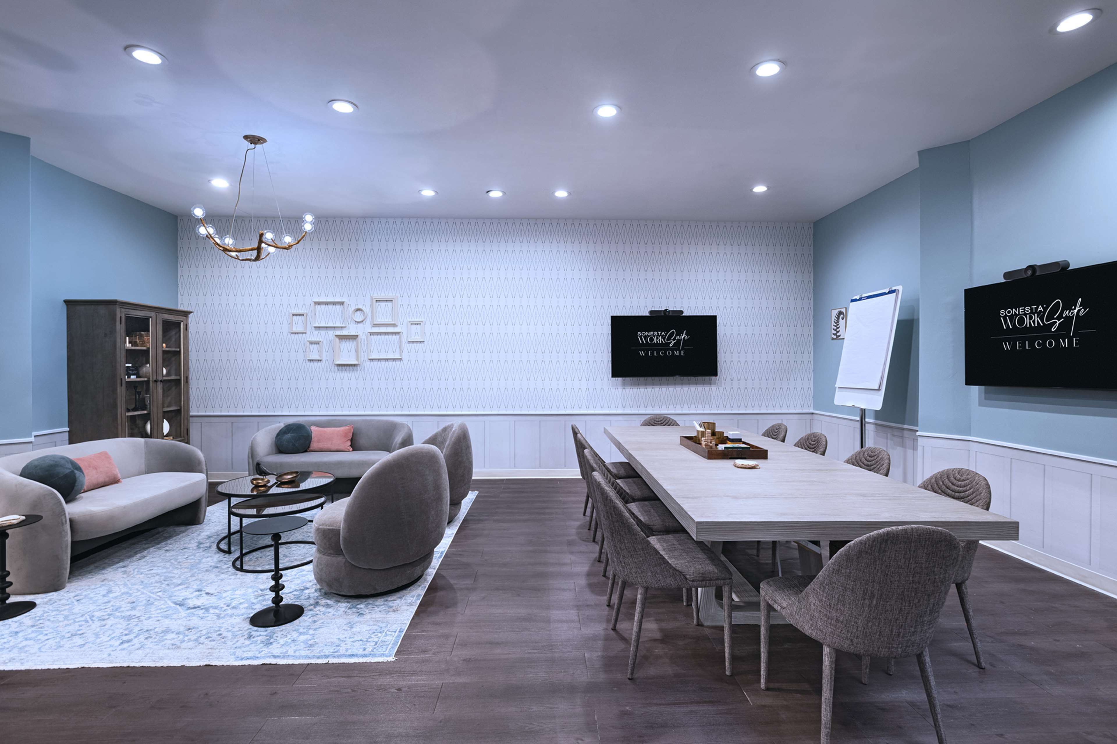 Una sala riunioni moderna con un lungo tavolo, sedute confortevoli, un lampadario e uno schermo TV che mostra un messaggio di benvenuto.
