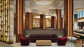 Un vestíbulo moderno con un sofá grande y capitonado, dos sillones y una mesa de centro de madera. Se observan columnas cilíndricas altas y una pared decorativa con un diseño geométrico.