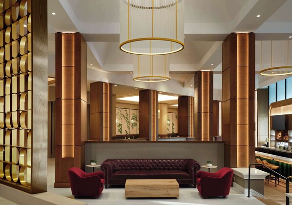 Un vestíbulo moderno con un sofá grande y capitonado, dos sillones y una mesa de centro de madera. Se observan columnas cilíndricas altas y una pared decorativa con un diseño geométrico.