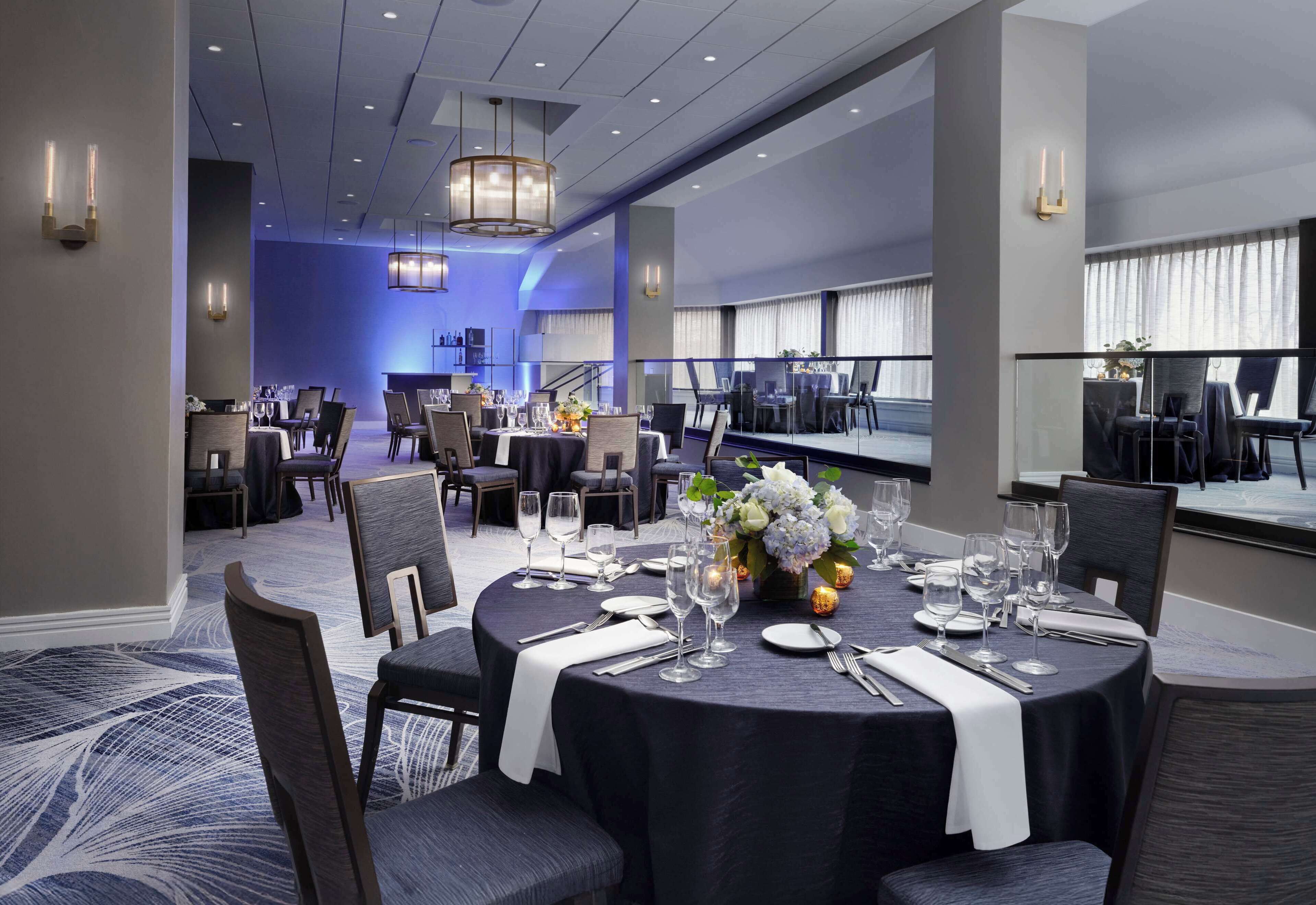 A dining area with round tables set for service, featuring a central floral arrangement and a modern chandelier.