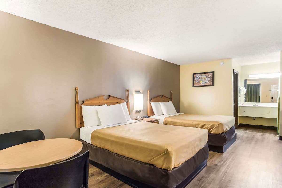 A hotel room with two beds, a round table, a chair, a bathroom with a mirror, and a wall-mounted picture.