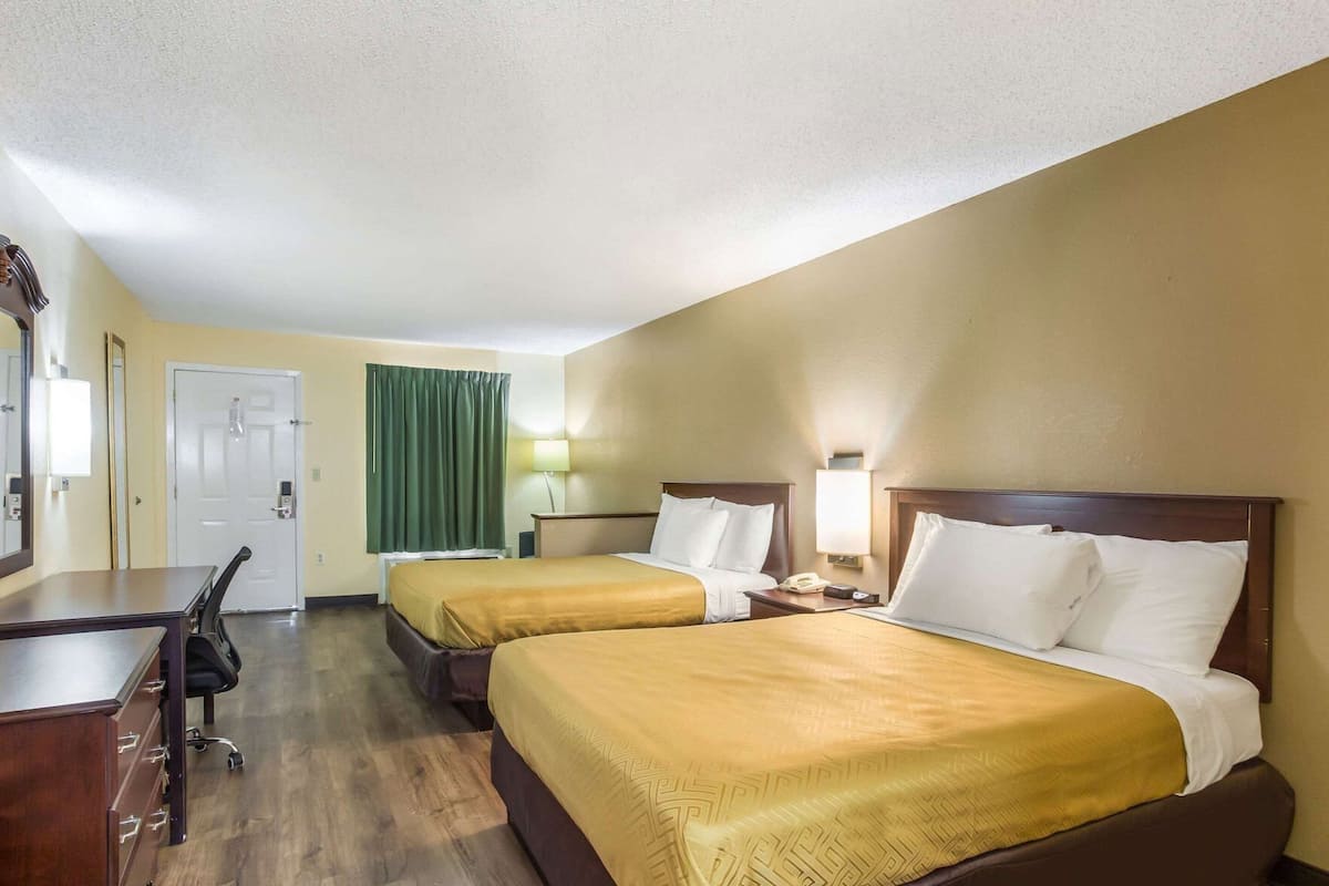 A hotel room with two beds, a desk, a chair, a lamp, and a door.