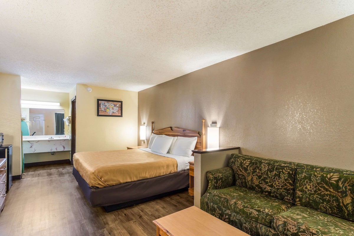 A hotel room with a bed, a green floral sofa, a wooden table, a desk with a mirror, and a wall-mounted lamp.