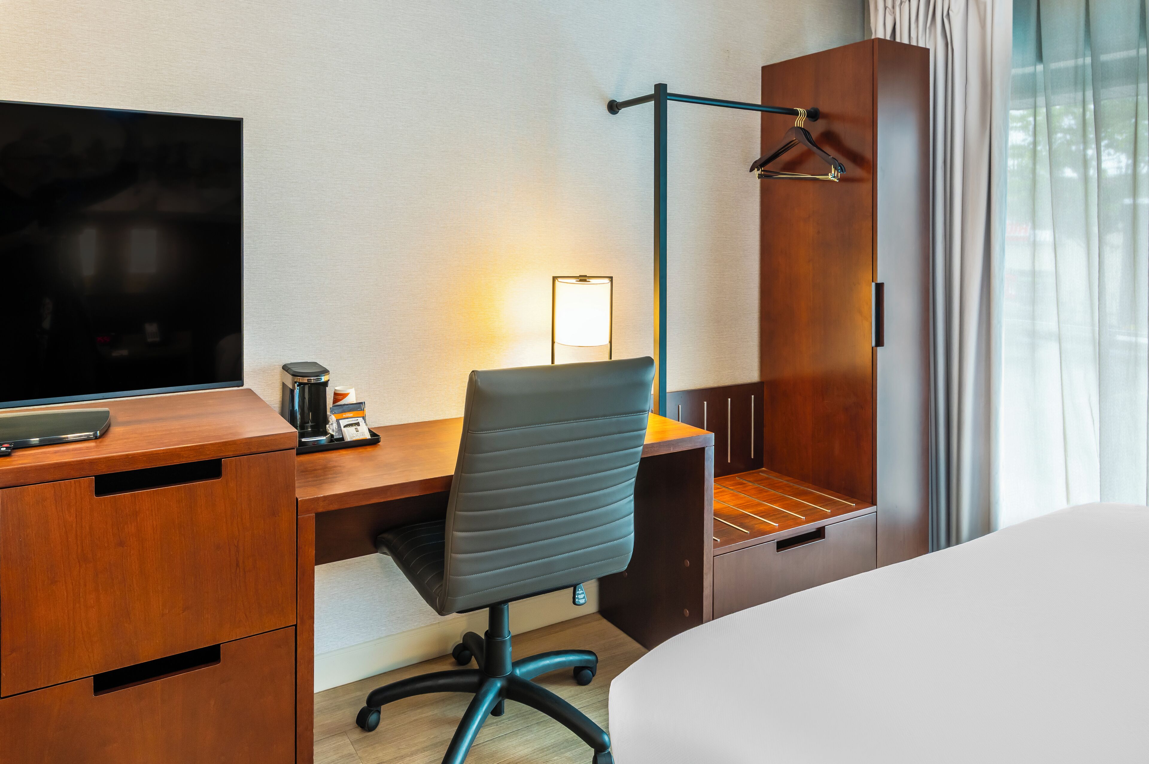 Een hotelkamer met een houten bureau, een zwarte bureaustoel, een televisie, een lamp en een kledingkast.