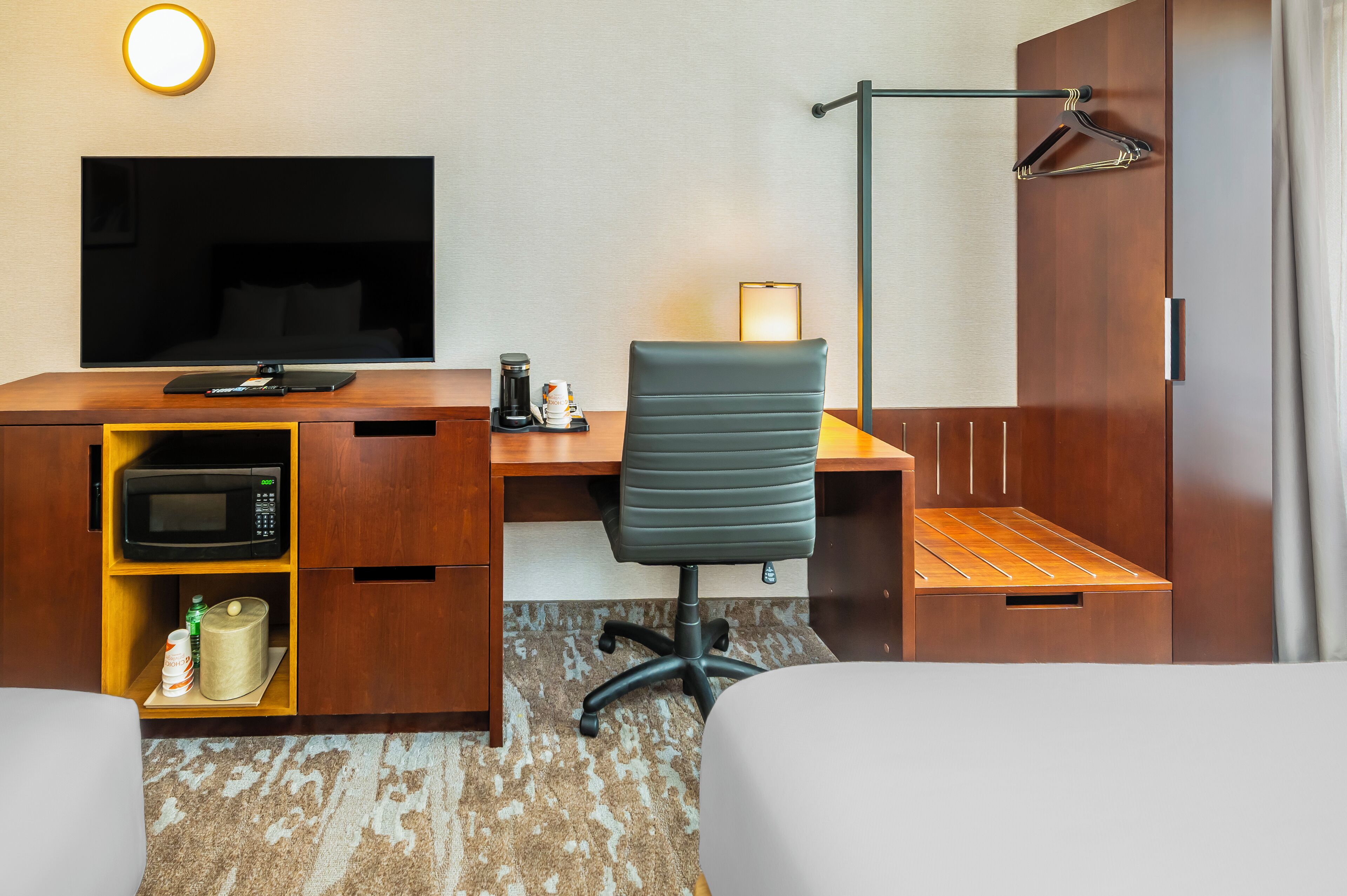 A hotel room with a flat-screen TV, a desk with a coffee maker, a chair, a bed, and a wardrobe.