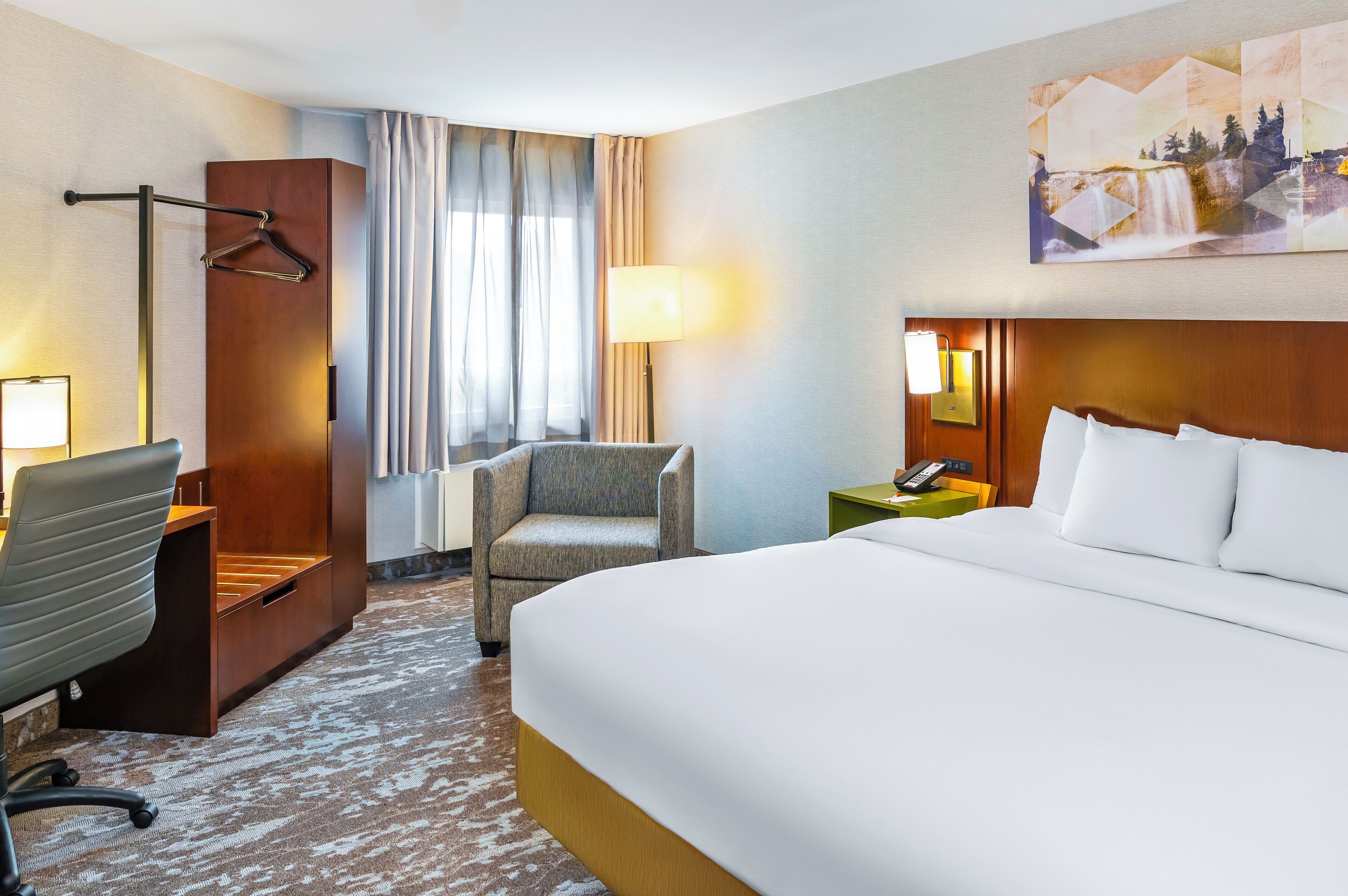 Habitación de hotel con cama, una silla, un escritorio, una lámpara y un cuadro en la pared.