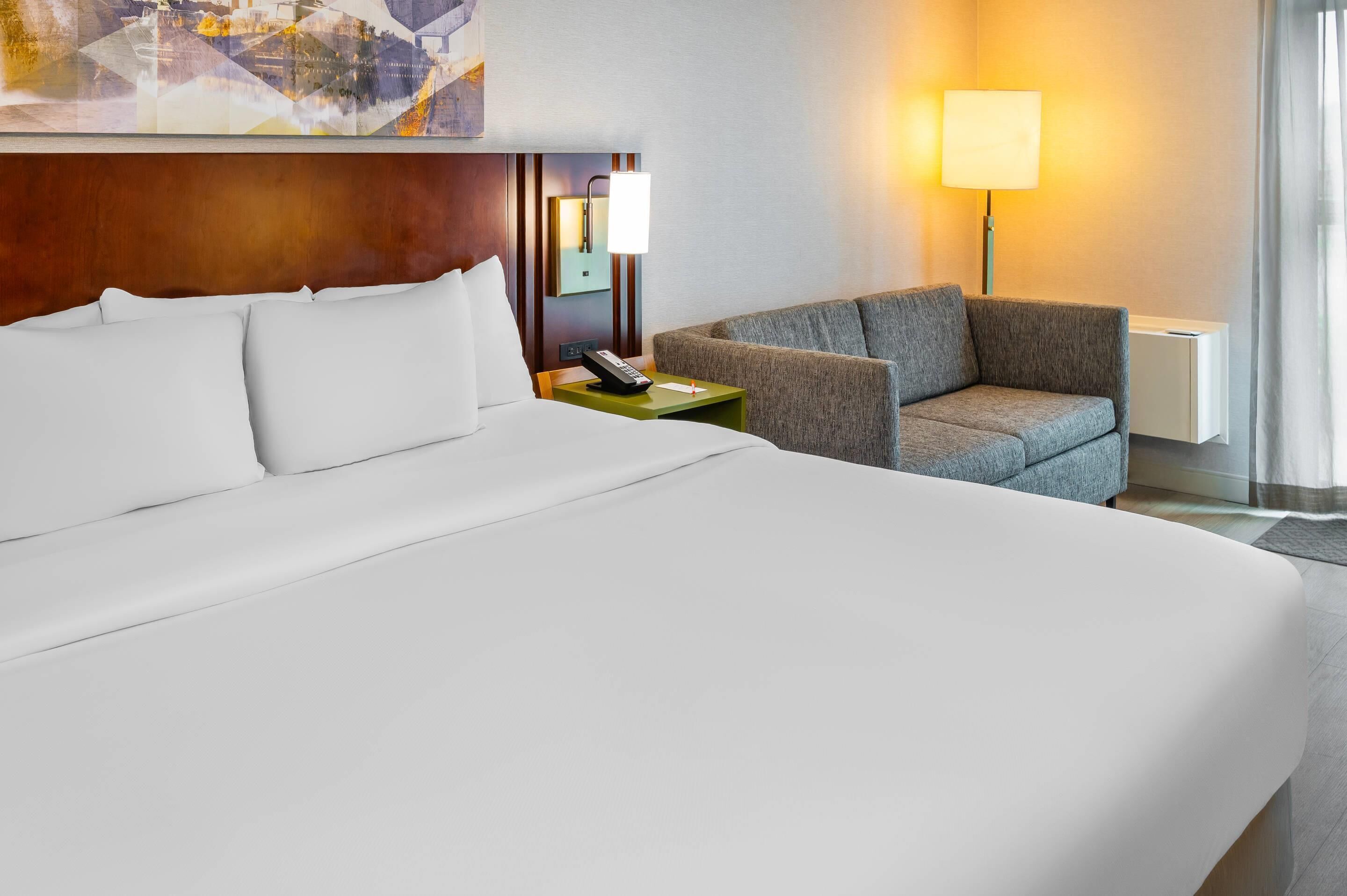 Een hotelkamer met een groot bed, een grijze fauteuil, een nachtkastje met een lamp en een schilderij aan de muur.