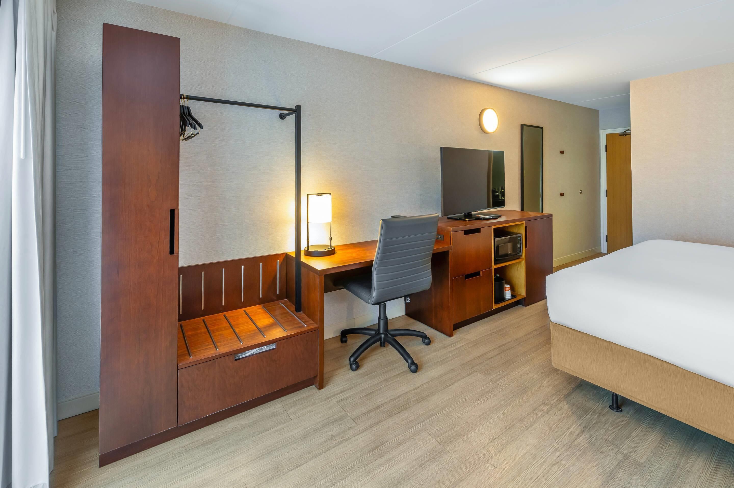 Et hotelværelse med seng, skrivebord med stol, garderobeskab, spejl og lampe.