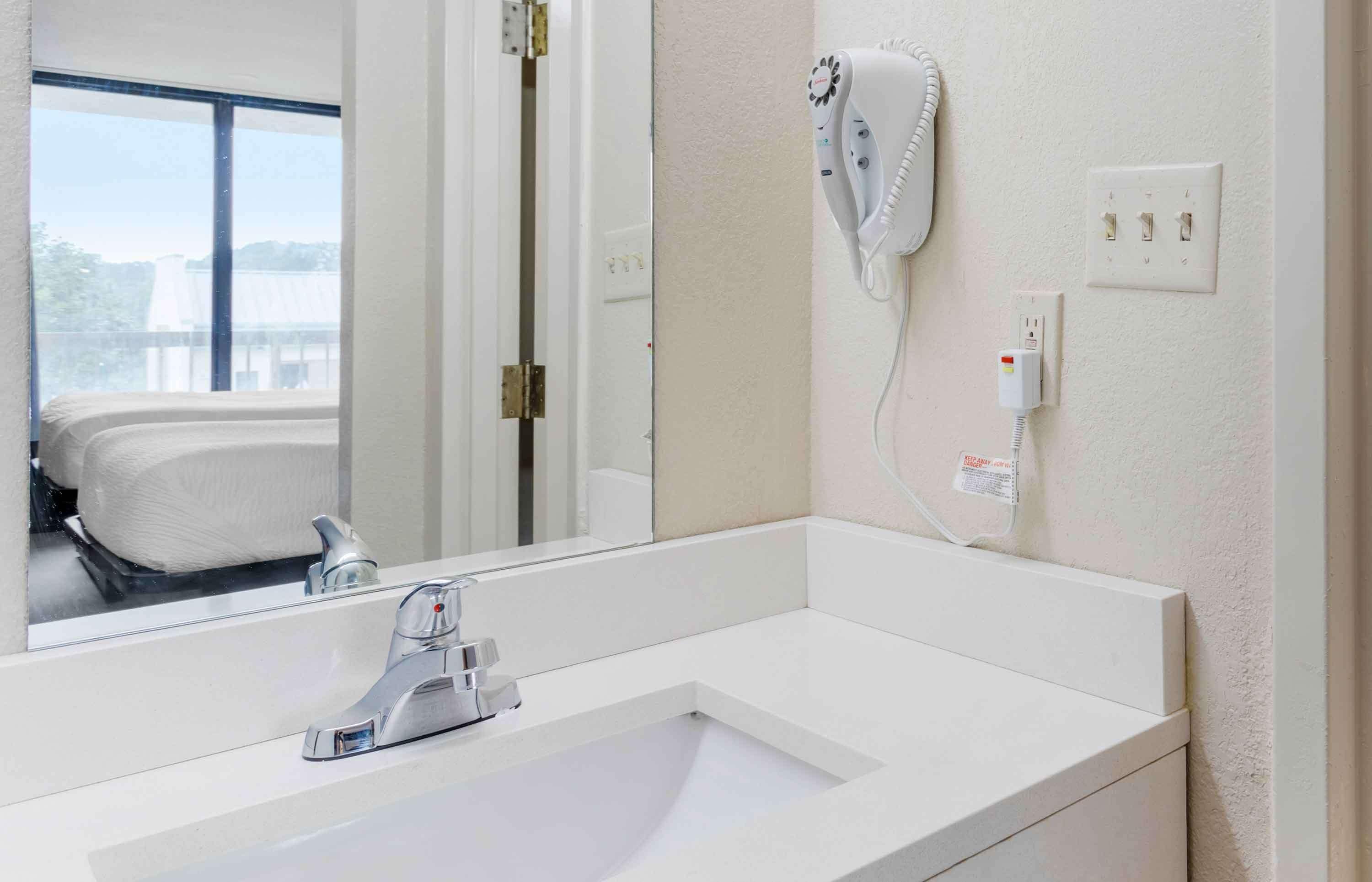 A modern bathroom with a large mirror, a white sink, a hairdryer mounted on the wall, and a window with a view of a building outside.