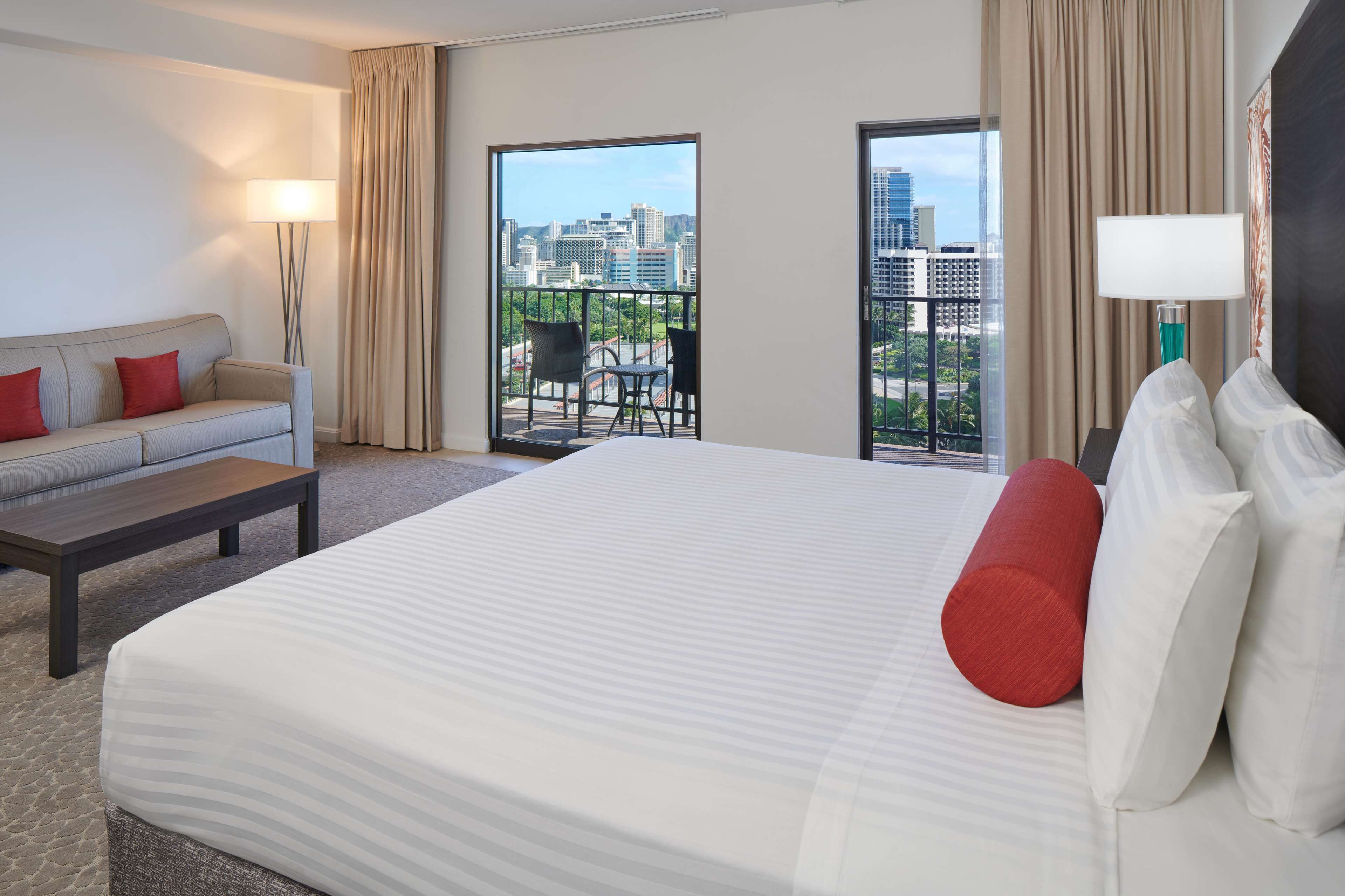 A hotel room with a large bed, a sofa, a coffee table, and a balcony with a city view.