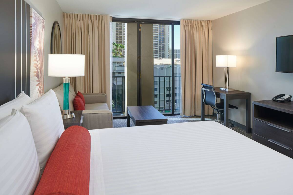 Quarto de hotel com uma cama grande, uma escrivaninha com cadeira, uma mesinha, televisão e varanda com cortinas.