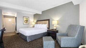 A hotel room with a bed, a chair, an ottoman, a desk, and a lamp.