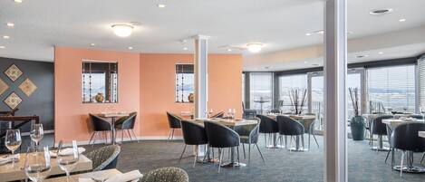 A hotel dining area with multiple tables and chairs, large windows, and a view of the city.