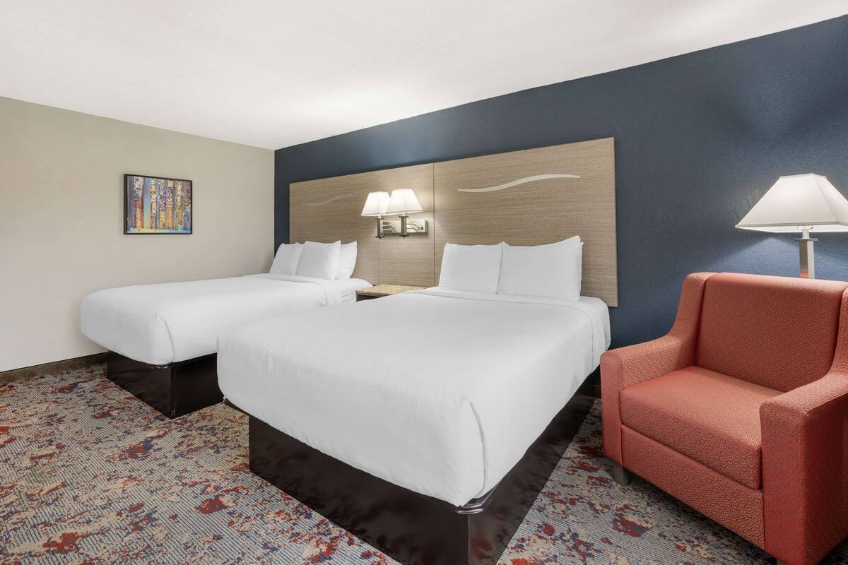 Quarto de hotel com duas camas, um sofá, cabeceira com luminárias, um quadro na parede e um tapete estampado.