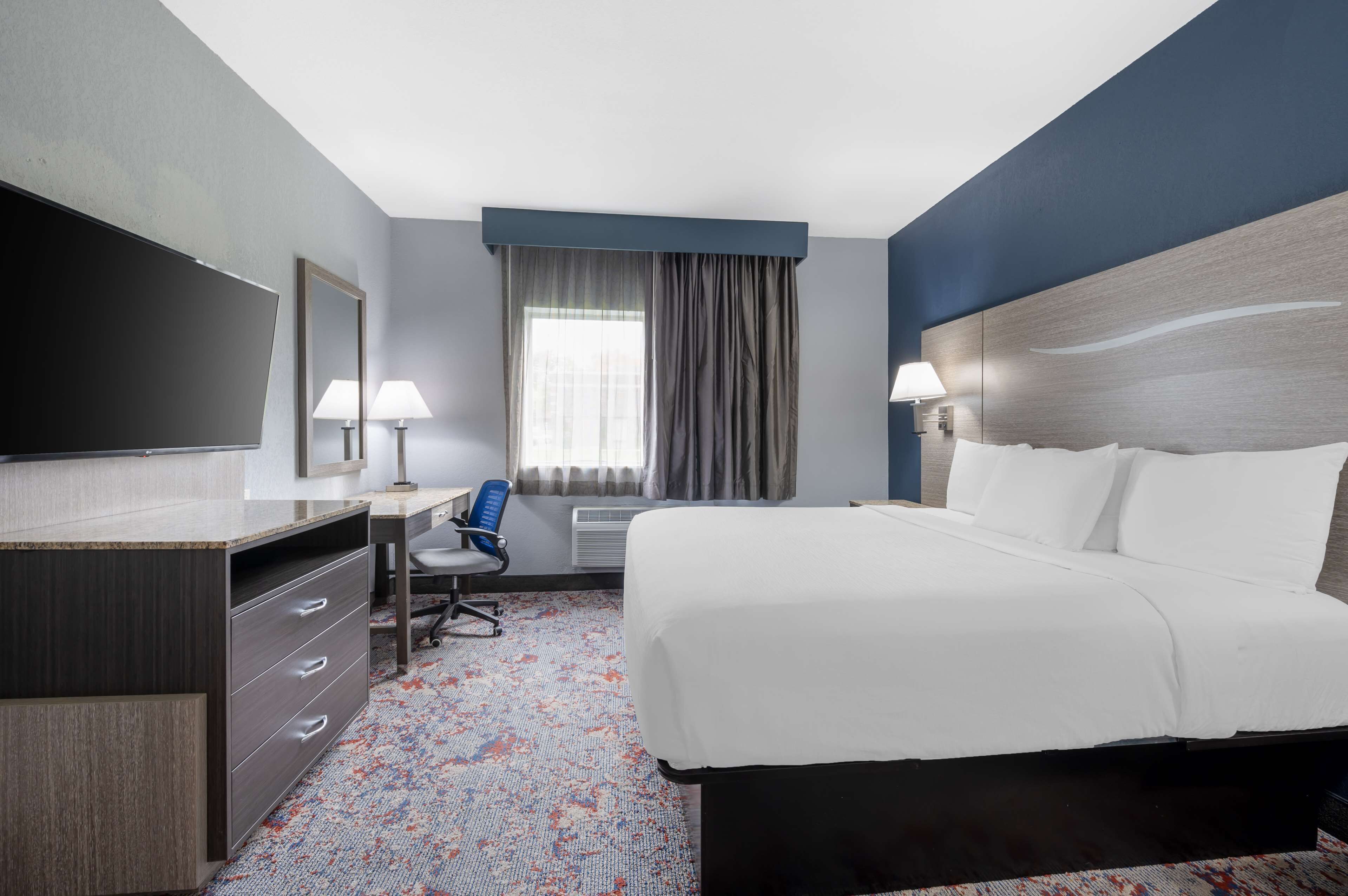 A hotel room with a large bed, a desk with a chair, a flat-screen TV, and a window with curtains.