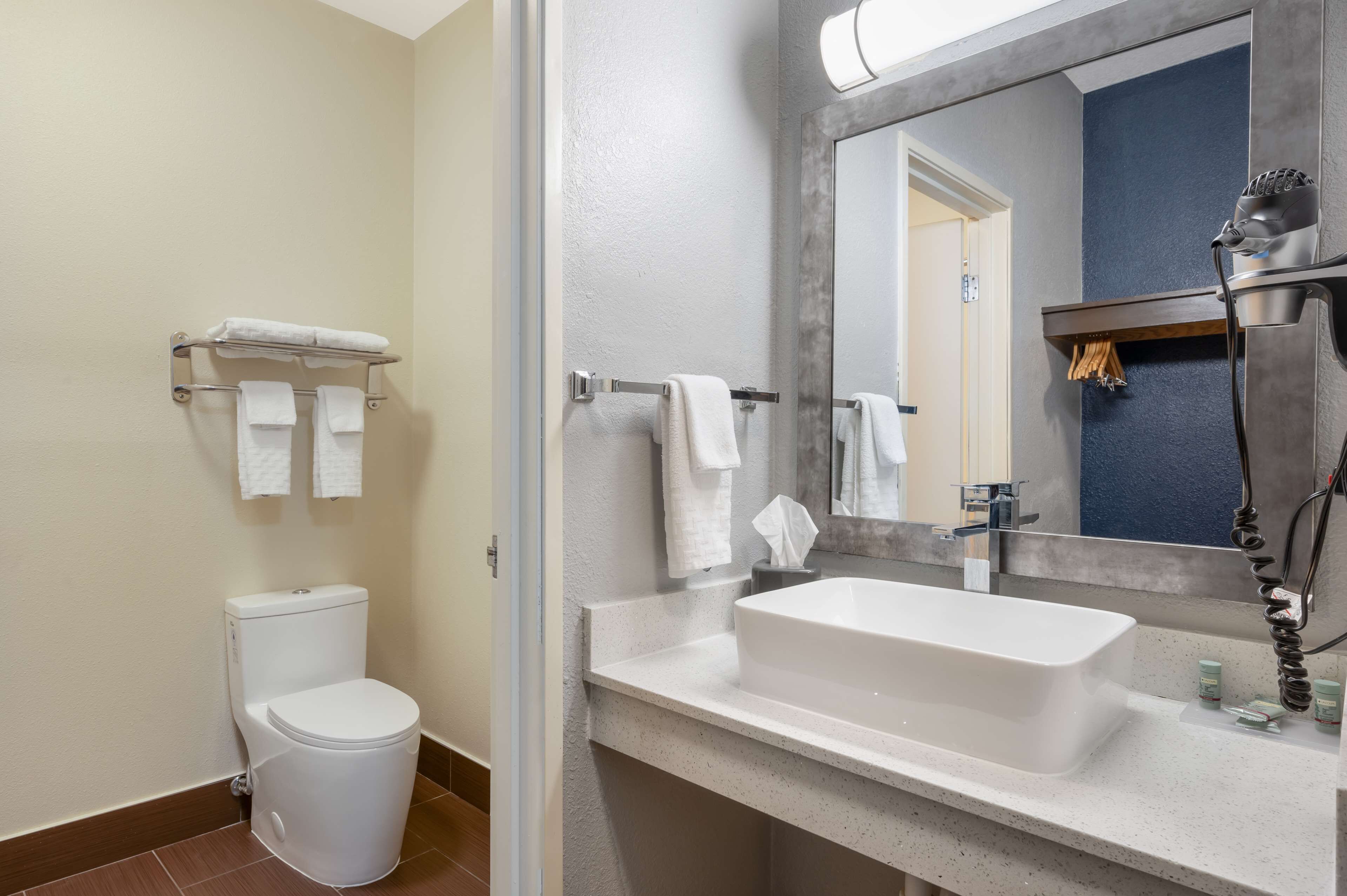 Un baño con inodoro blanco, un lavabo con un espejo grande y un perchero con varias toallas.