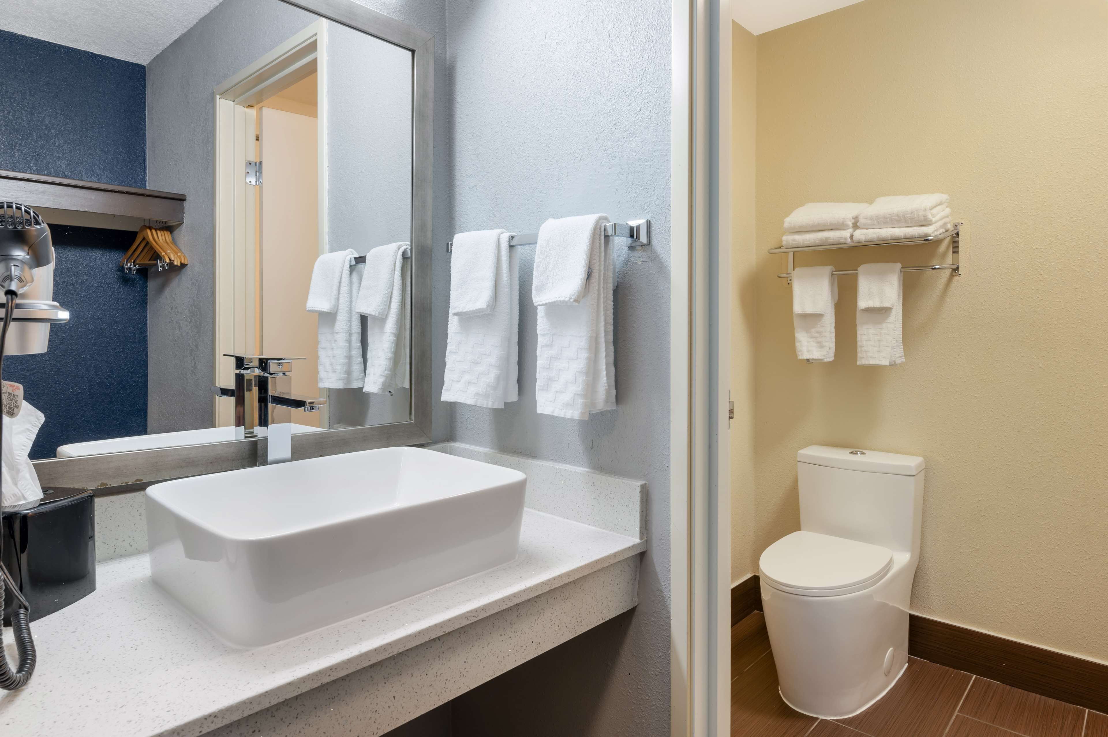 A modern bathroom with a large white sink, a mirror, and a toilet. There are multiple white towels hanging on a rack.