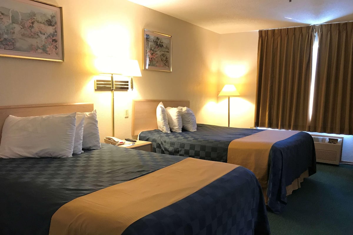 A hotel room with two beds, a nightstand, a lamp, a painting, and a window with curtains.