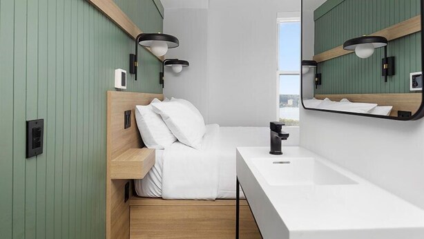 A modern hotel room with a wooden headboard, a white bed, a large mirror, and a white sink. - Riverside Tower Hotel (New York)