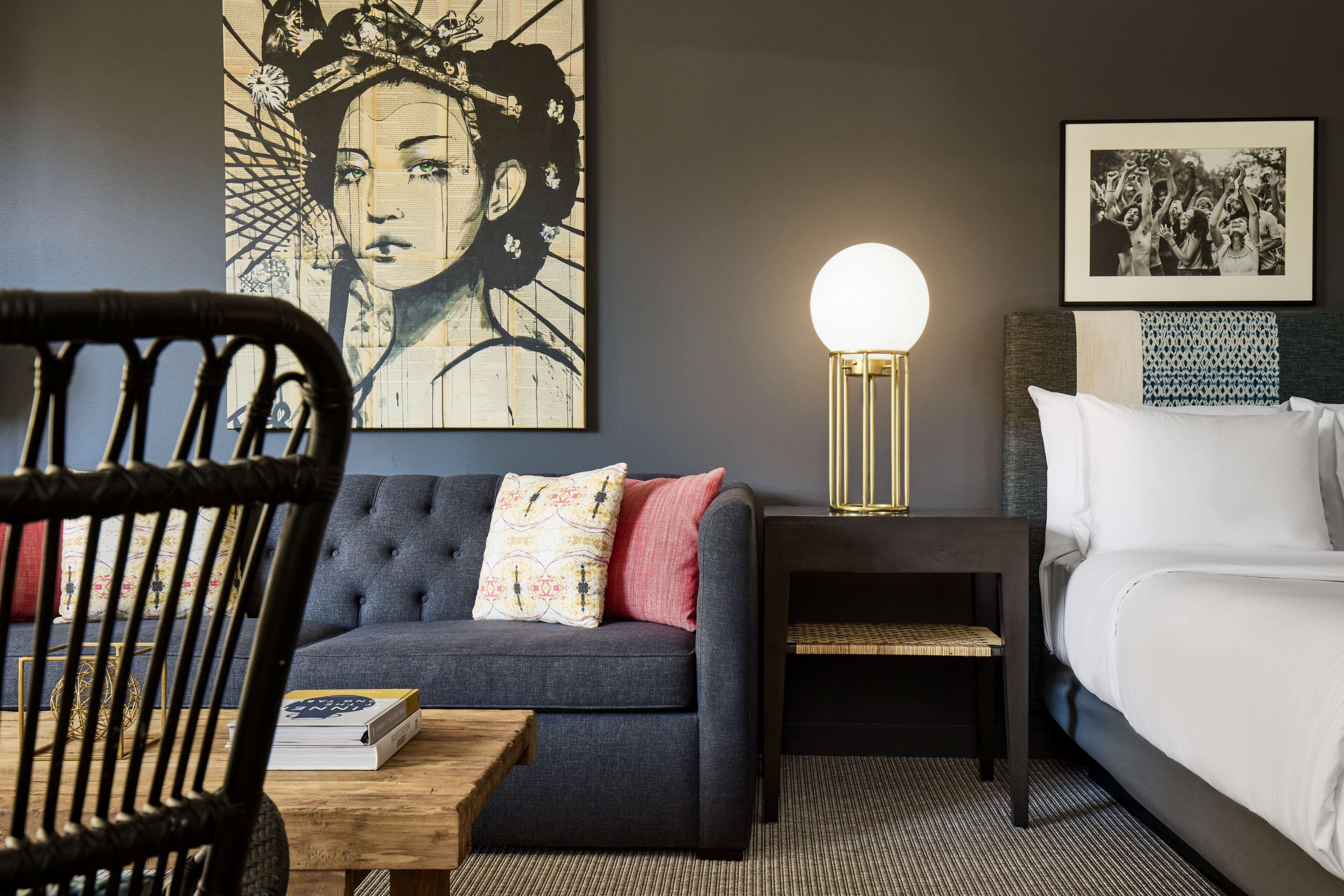 Una habitación de hotel moderna con un sofá gris oscuro, una mesa de centro de madera, una mesita de noche con lámpara, una cama con ropa blanca y obras de arte en las paredes.