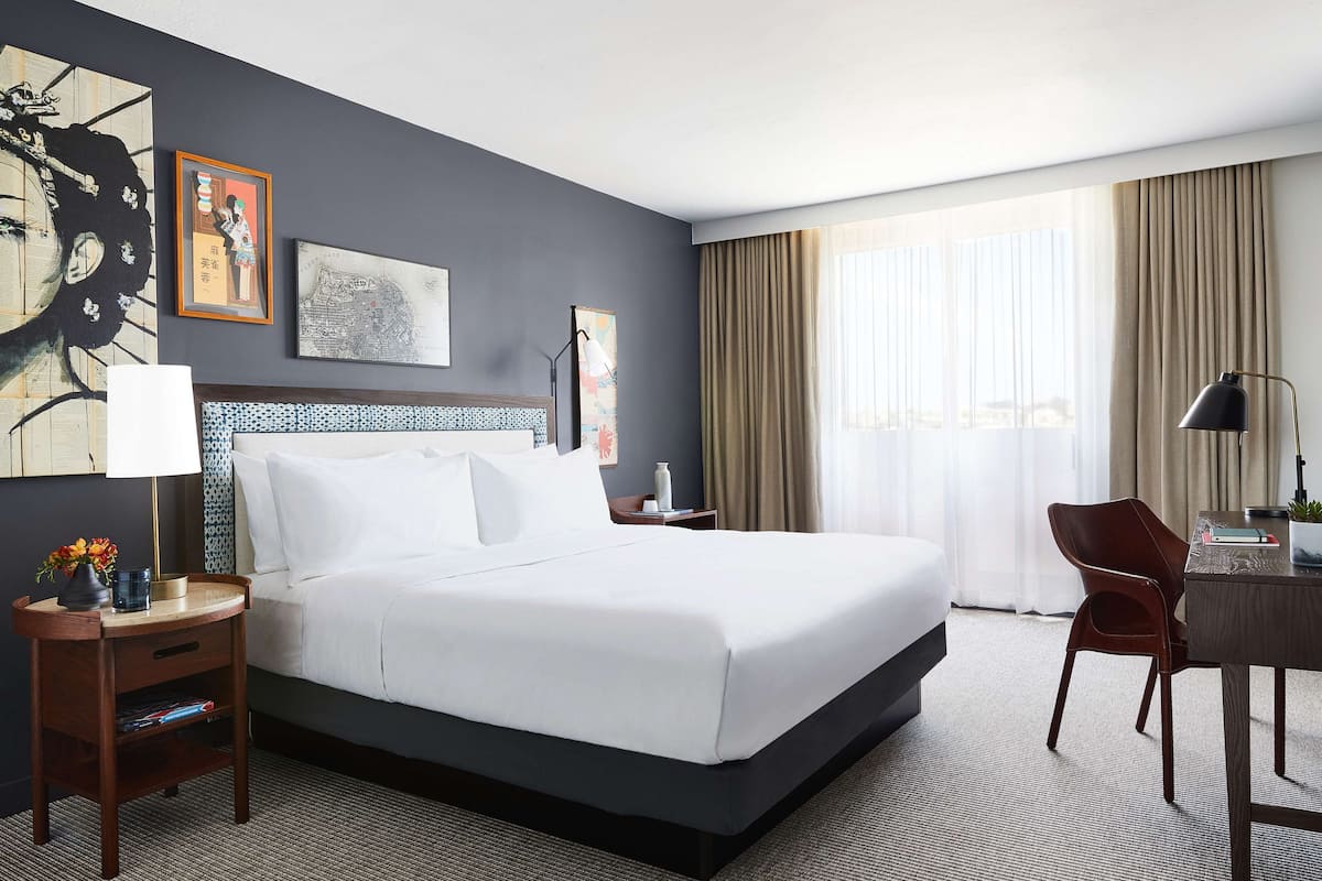 Ein Hotelzimmer mit einem großen Bett, einem Schreibtisch, einem Sessel und Kunstwerken an den Wänden.