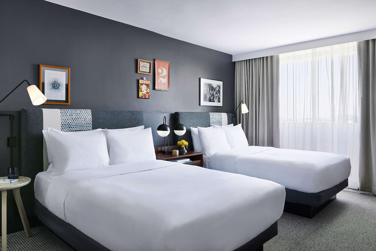 Ein Hotelzimmer mit zwei Betten, einem Nachttisch mit Lampe, einem kleinen Tisch mit einem Glas und einem großen Fenster mit Vorhängen.