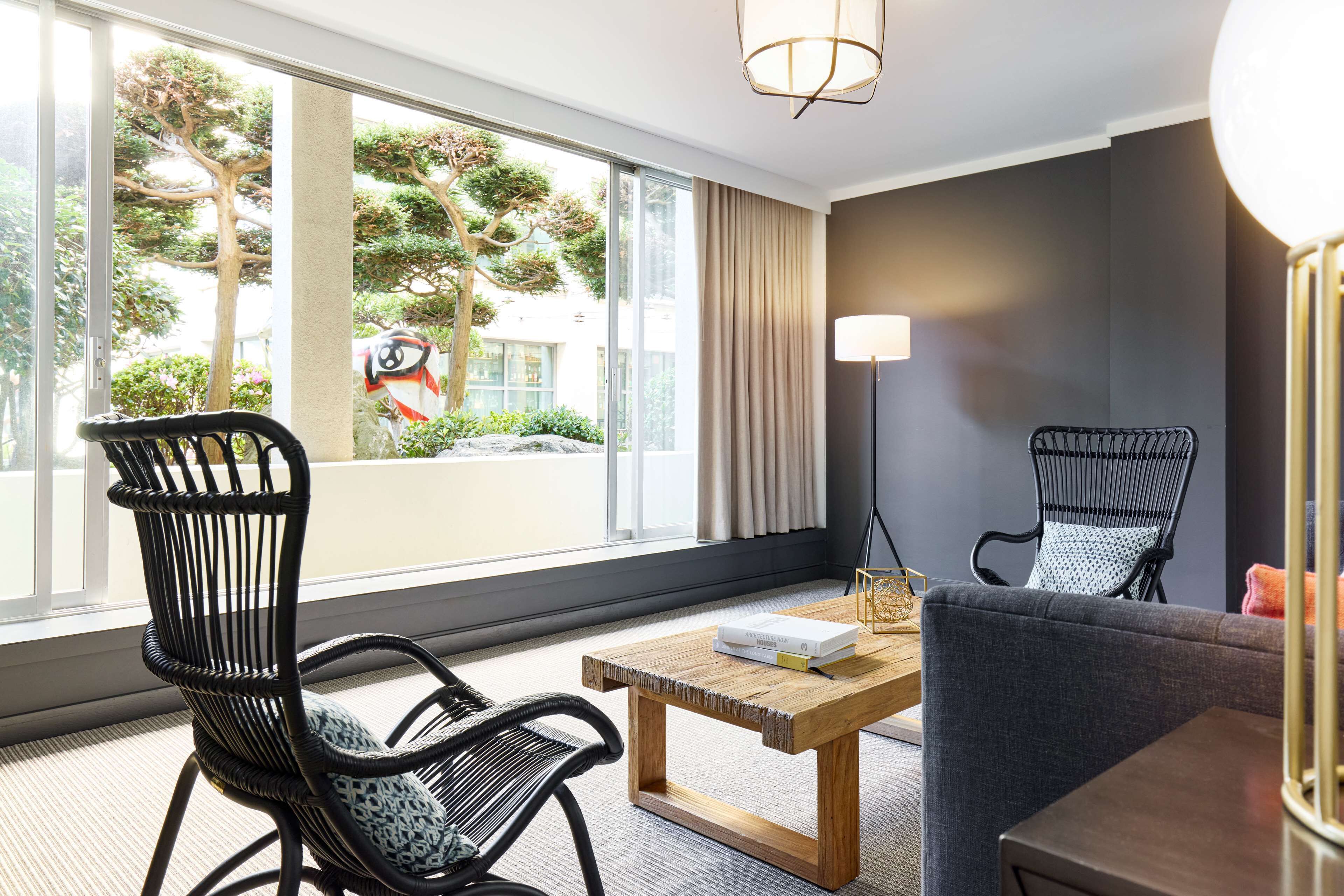 Habitación de hotel moderna con un gran ventanal, dos sillas de mimbre negro, un sofá gris, una mesa de centro de madera y una lámpara de pie.