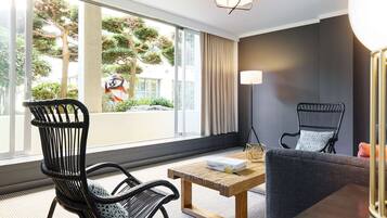 Habitación de hotel moderna con un gran ventanal, dos sillas de mimbre negro, un sofá gris, una mesa de centro de madera y una lámpara de pie.