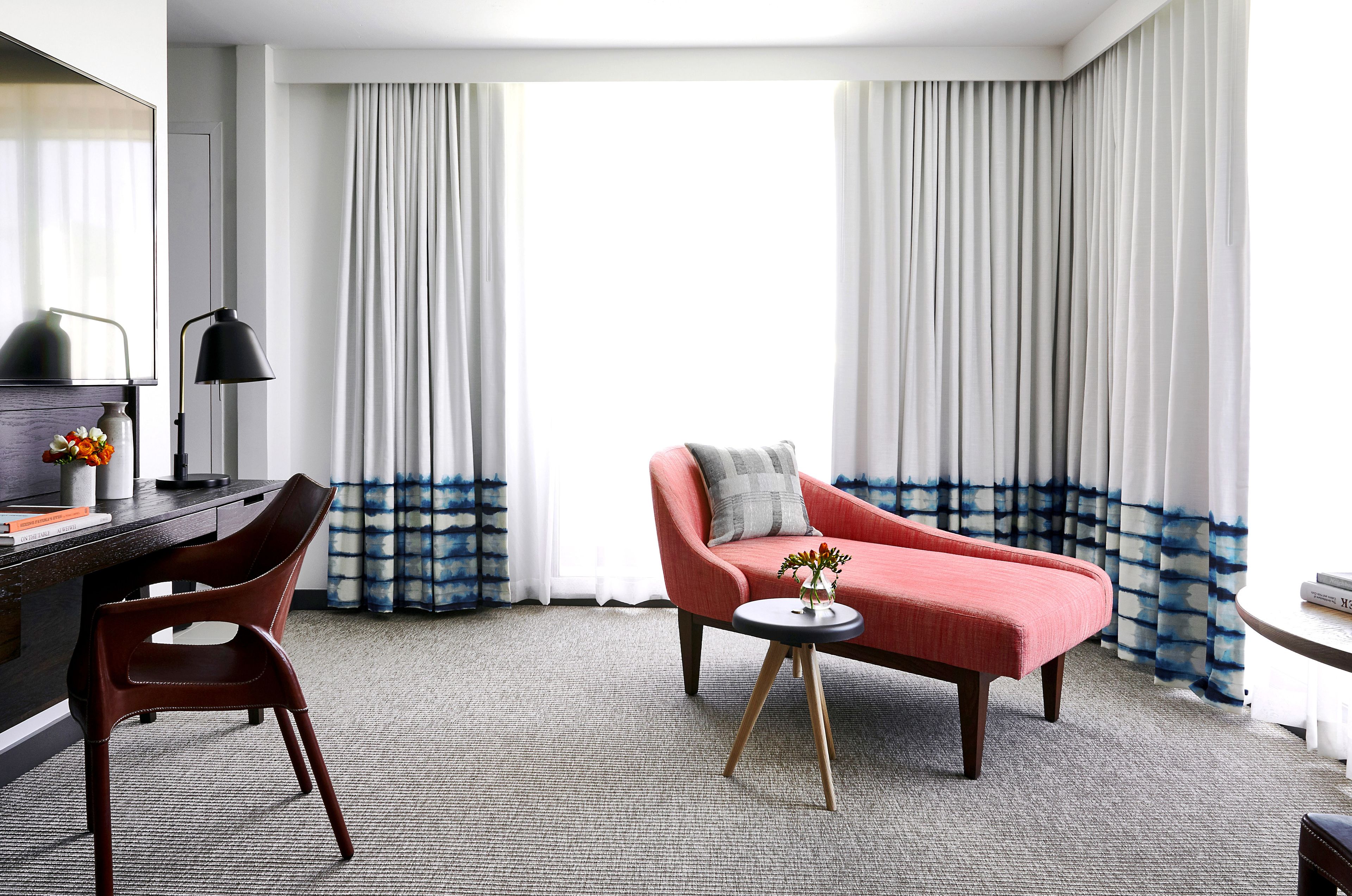 Habitación de hotel con sofá rojo, una silla de madera, una mesita redonda con un florero y un escritorio con lámpara.