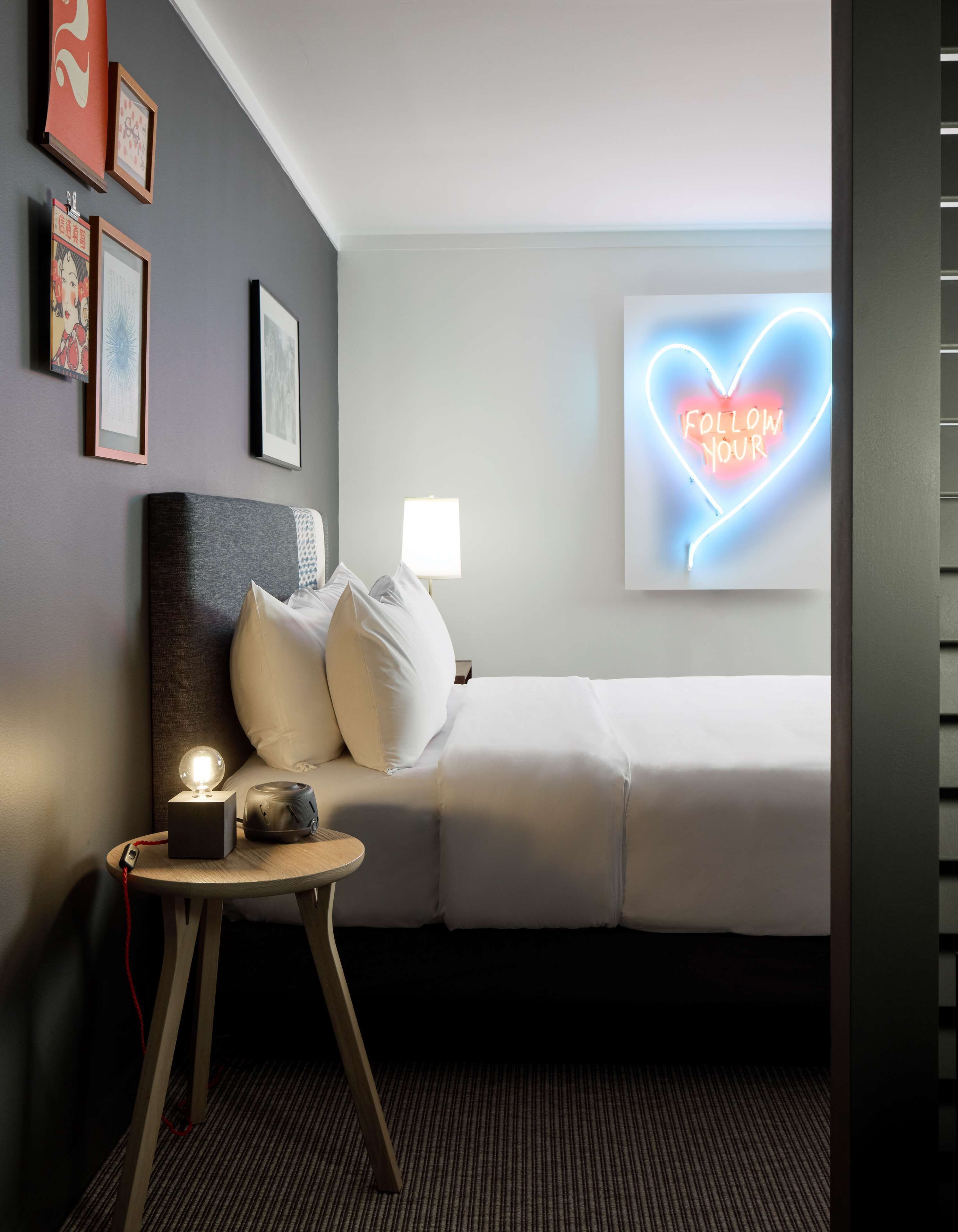 Habitación de hotel con cama, mesita de noche con lámpara, letrero de neón y cuadros en la pared.