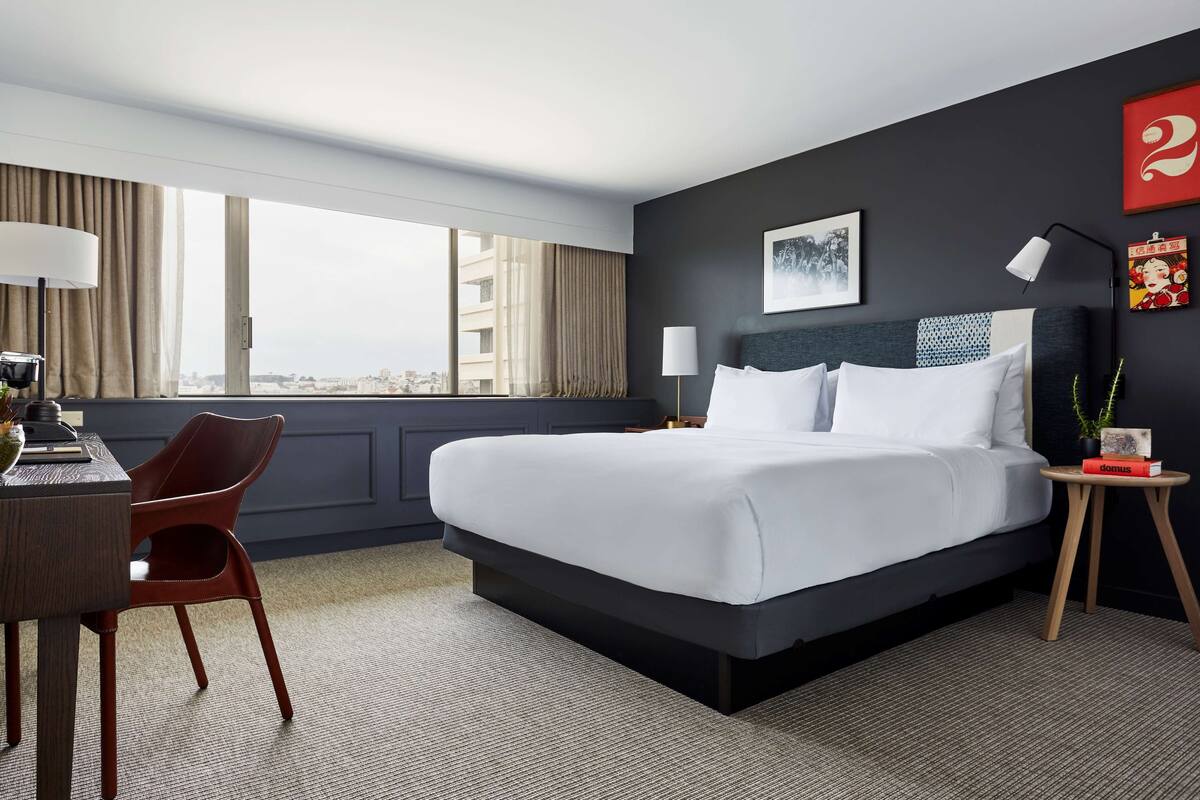 Ein Hotelzimmer mit einem großen Bett, einem Schreibtisch mit Stuhl, einem kleinen Beistelltisch und Blick auf die Stadt durch große Fenster.
