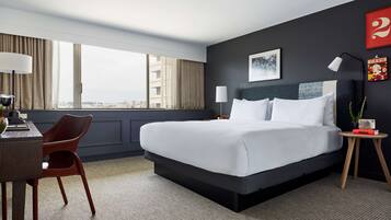 A hotel room with a large bed, a desk with a chair, a small side table, and a view of the city through large windows.
