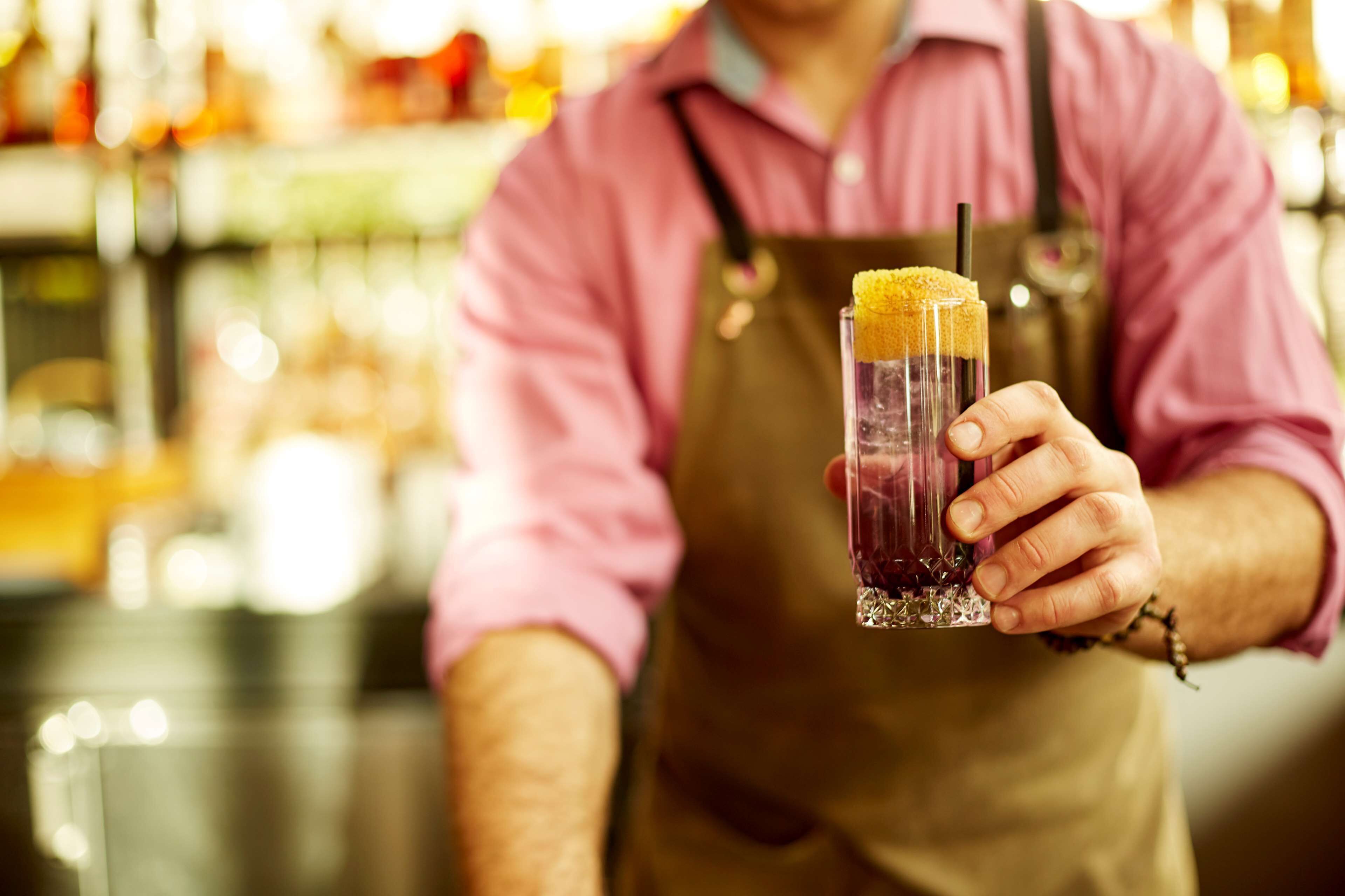 A person holding a tall glass with a straw and a yellow garnish, wearing a pink shirt and a brown apron.