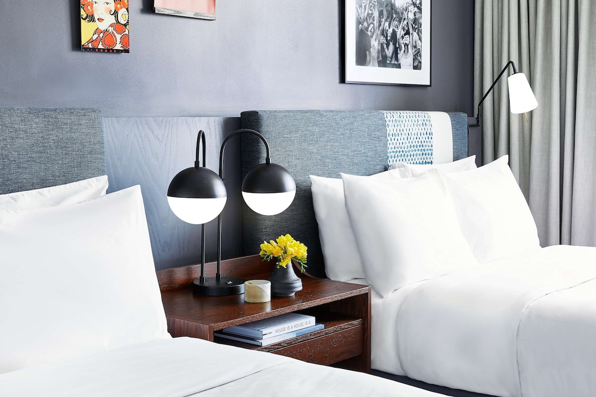 Ein Hotelzimmer mit einem ordentlich bezogenem Bett, einem Nachttisch mit zwei schwarzen Lampen, einer kleinen Vase mit gelben Blumen und einem gerahmten Bild an der Wand.