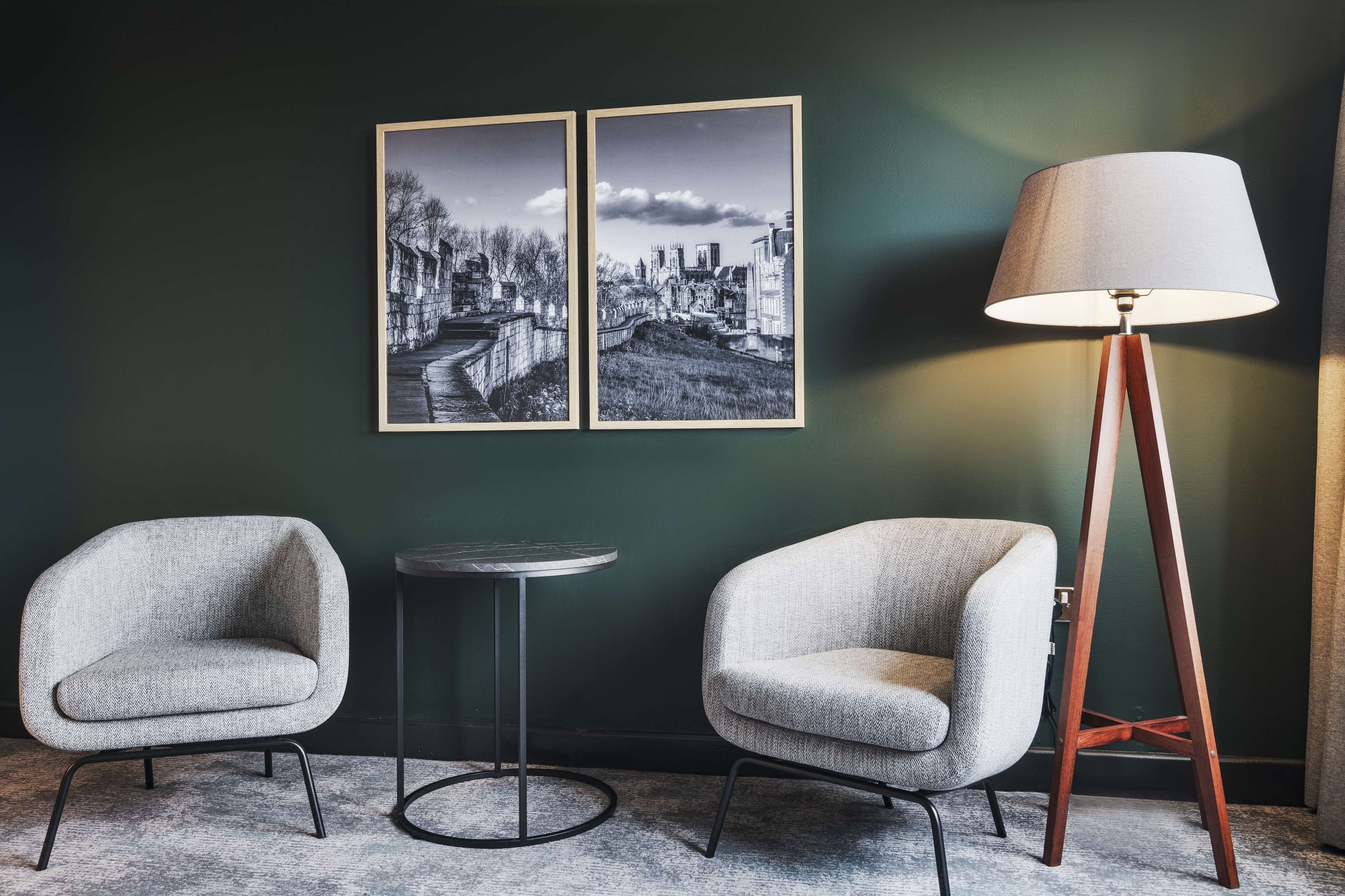 Une pièce avec deux fauteuils, une petite table ronde et un lampadaire placé contre un mur vert foncé. Deux photographies en noir et blanc sont accrochées au-dessus des fauteuils.