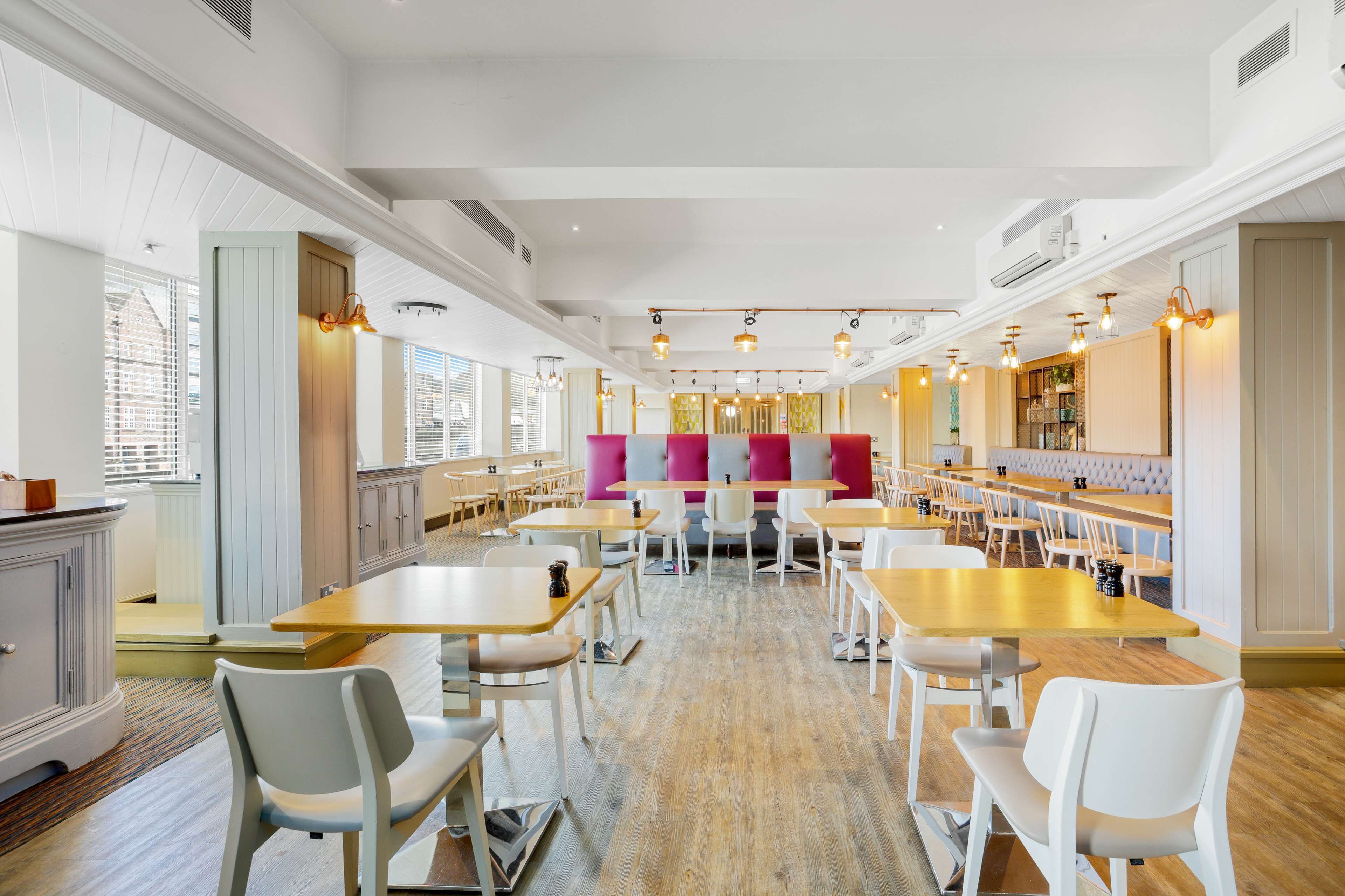 A spacious dining area with multiple tables and chairs, a central seating area with a pink and gray color scheme, and large windows providing natural light.
