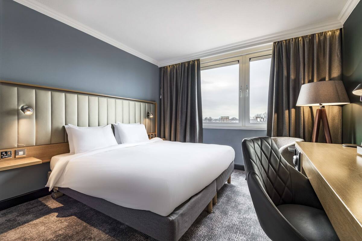 A hotel room with a large bed, a seating area, a desk, and a window with curtains.