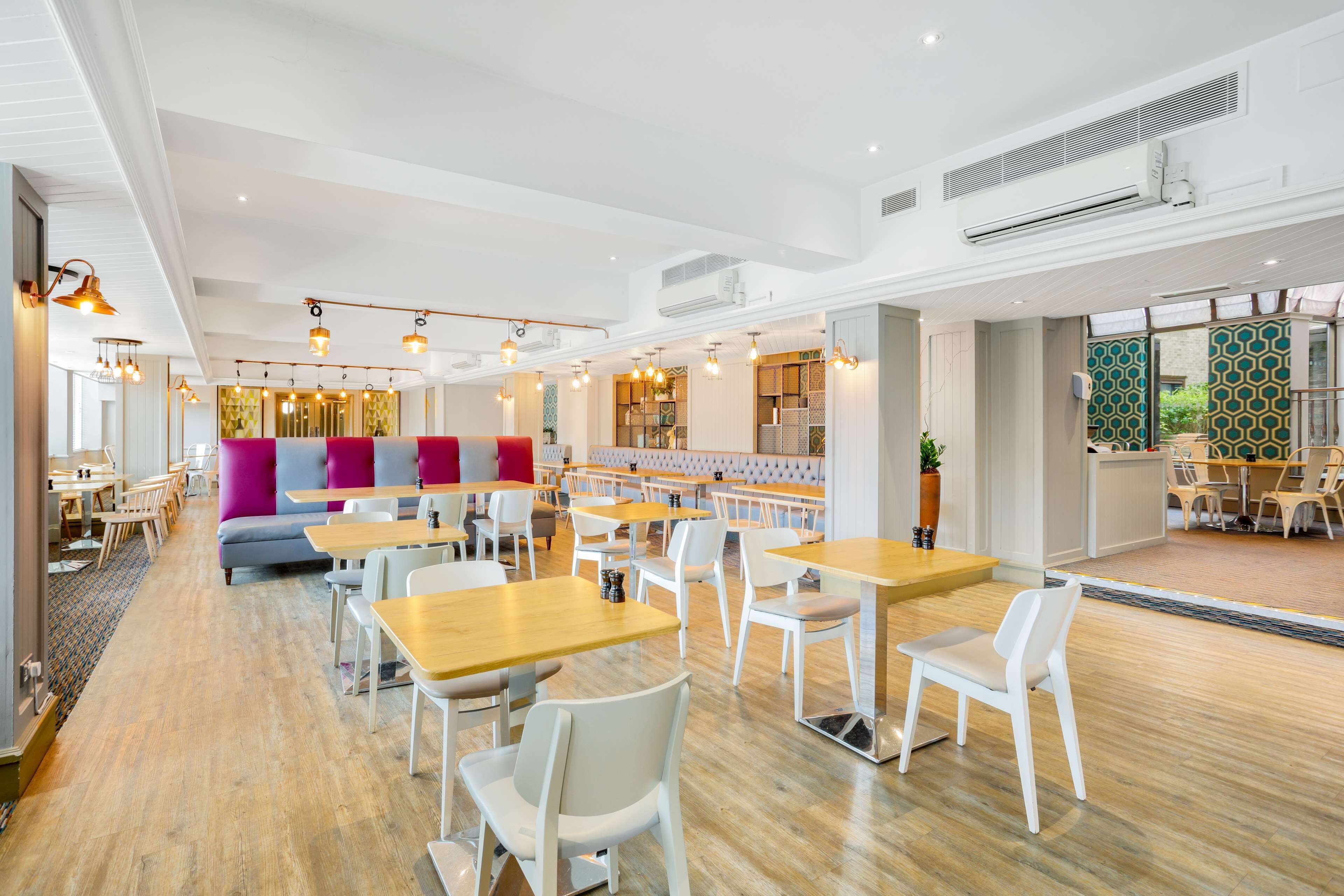 A modern dining area with wooden tables and white chairs, a colorful seating area, and a view of the outdoors.