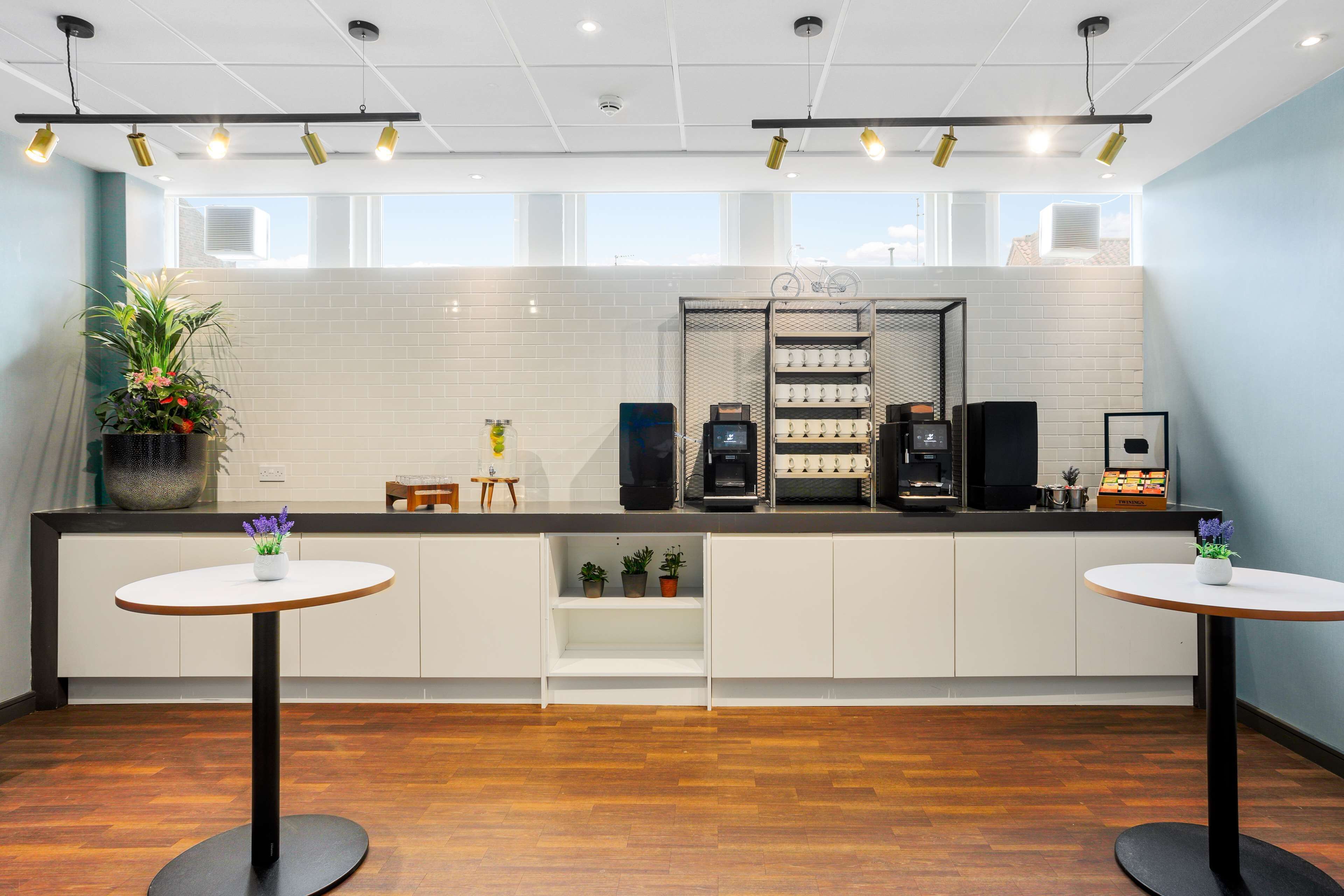 A modern kitchen with white cabinets, a black countertop, and a coffee machine. There are two small round tables with white tops and black bases.
