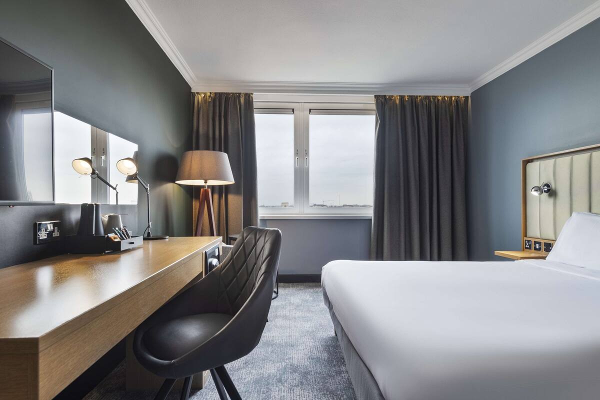 A hotel room with a bed, a desk with a chair, a lamp, a mirror, and a window with curtains.