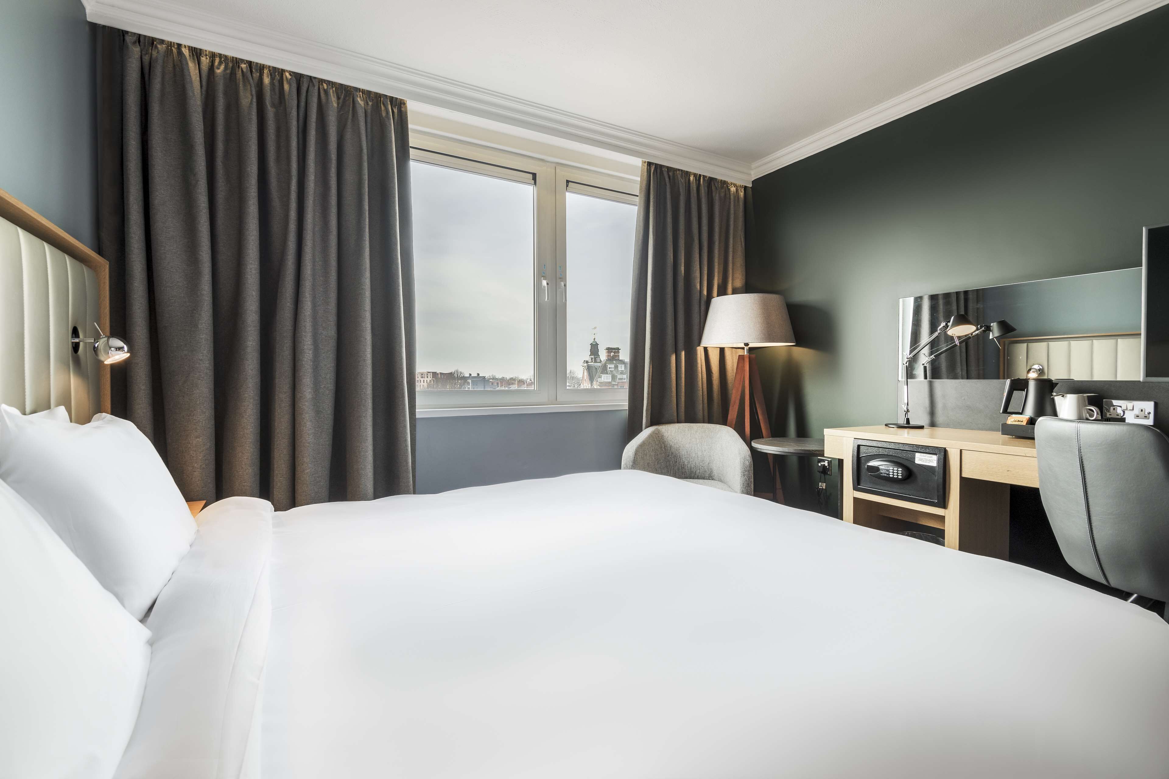 A hotel room with a large bed, a desk with a lamp, a chair, and a view of a cityscape through the window.