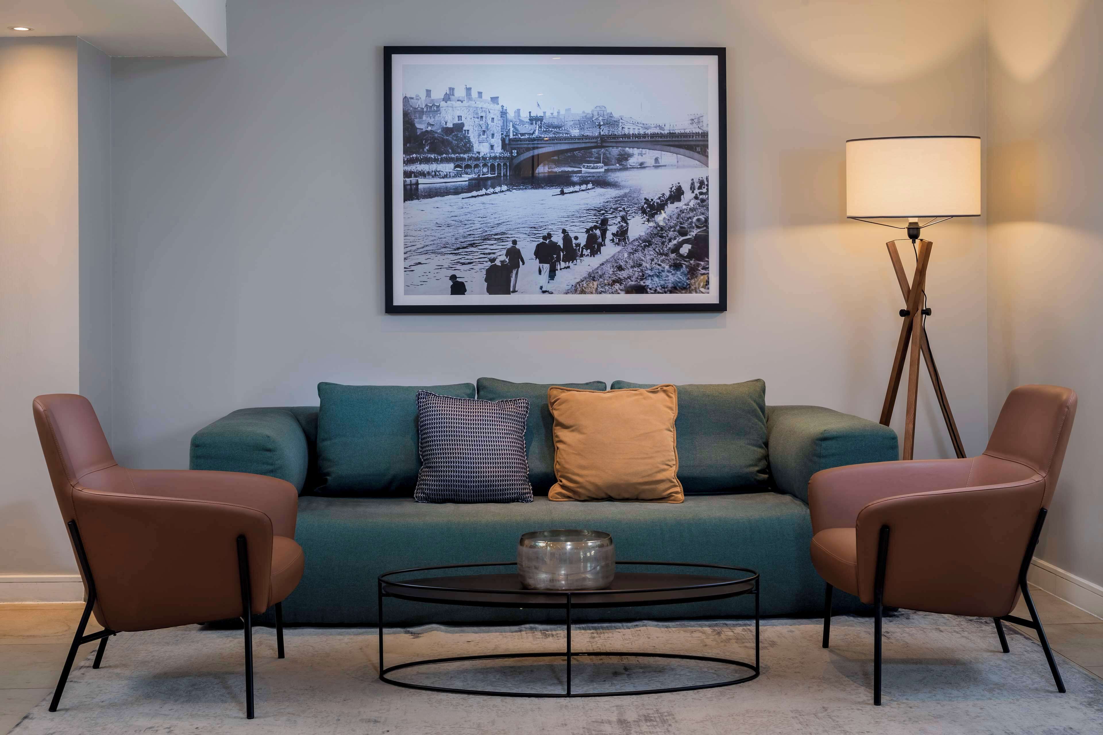 A living room with a teal sofa, two brown armchairs, a round coffee table, and a floor lamp. A framed black and white photograph of a bridge and river is on the wall.