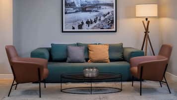A living room with a teal sofa, two brown armchairs, a round coffee table, and a floor lamp. A framed black and white photograph of a bridge and river is on the wall.