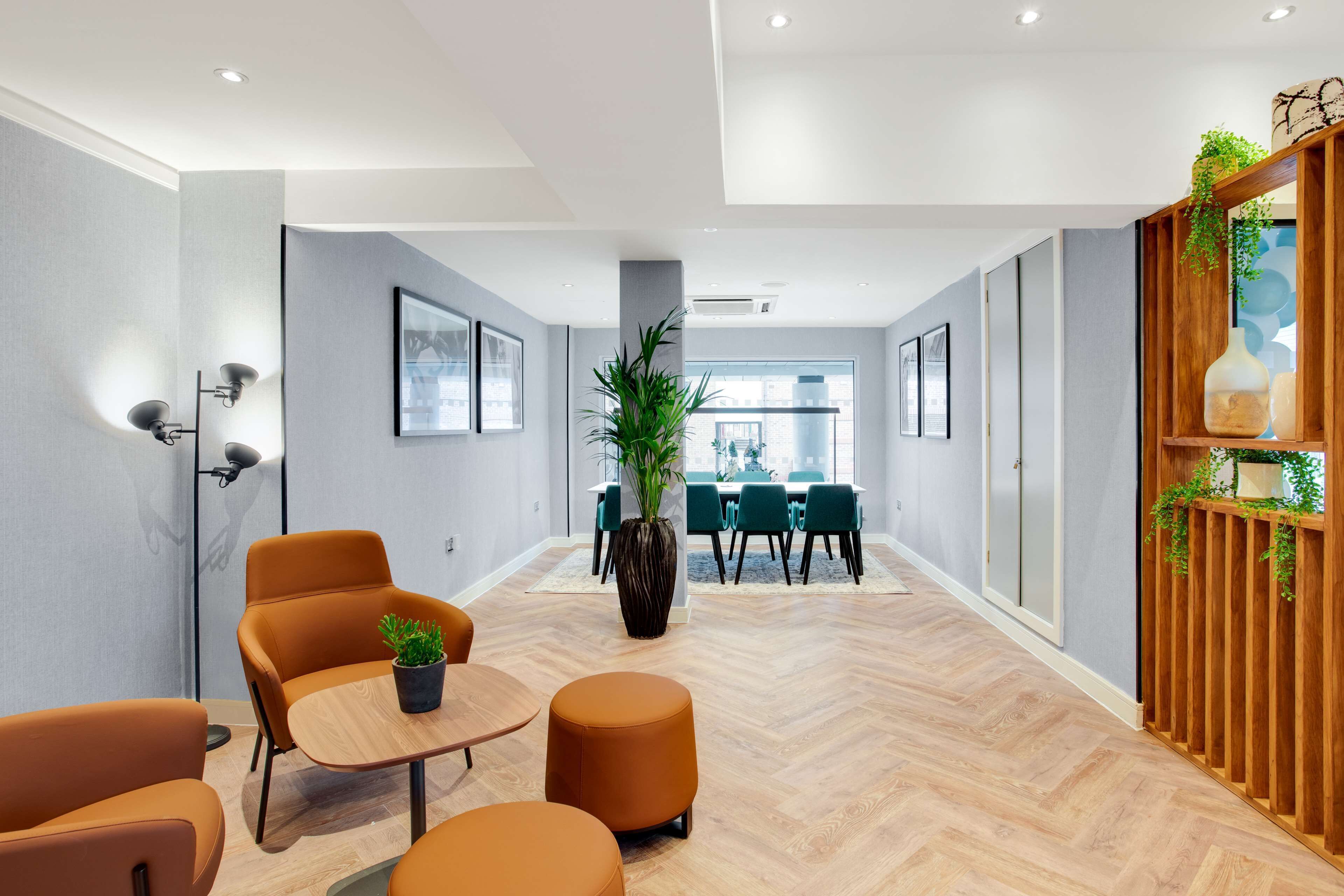 Un interior moderno con suelo de madera en espiga, una planta central, asientos naranjas y una zona de comedor con sillas verdes.