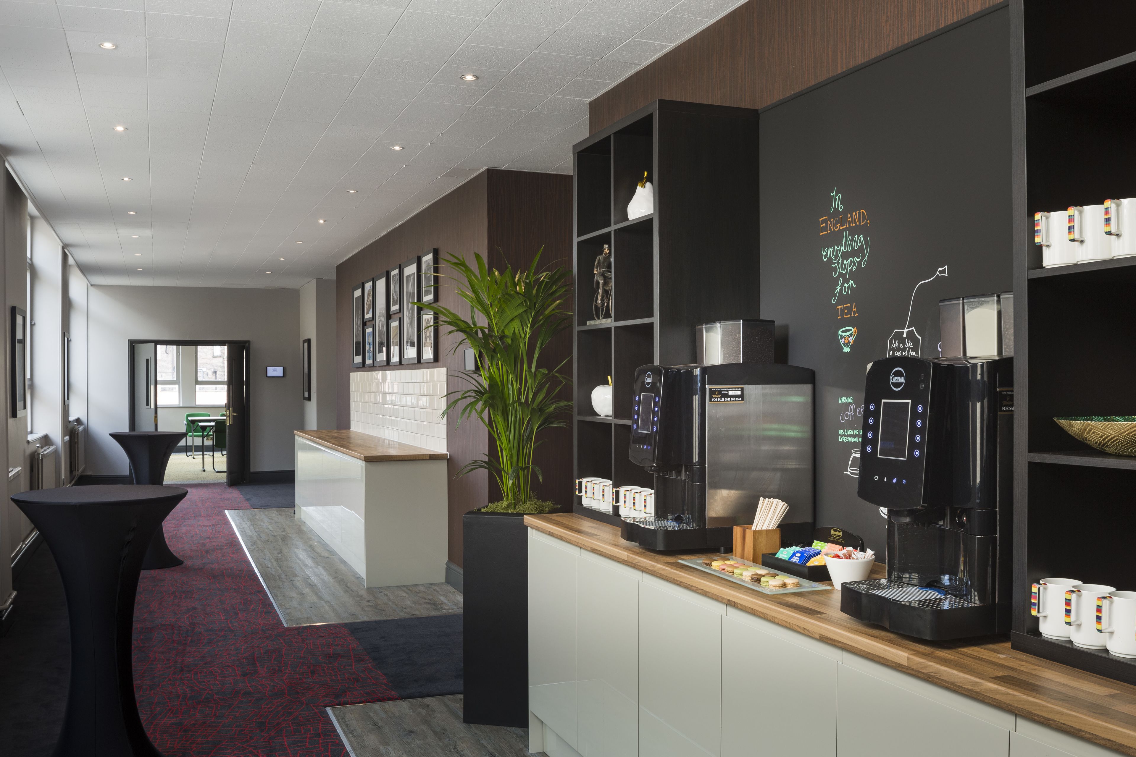 Un moderno vestíbulo de hotel con un mostrador de recepción, máquinas de café y una planta grande en maceta.